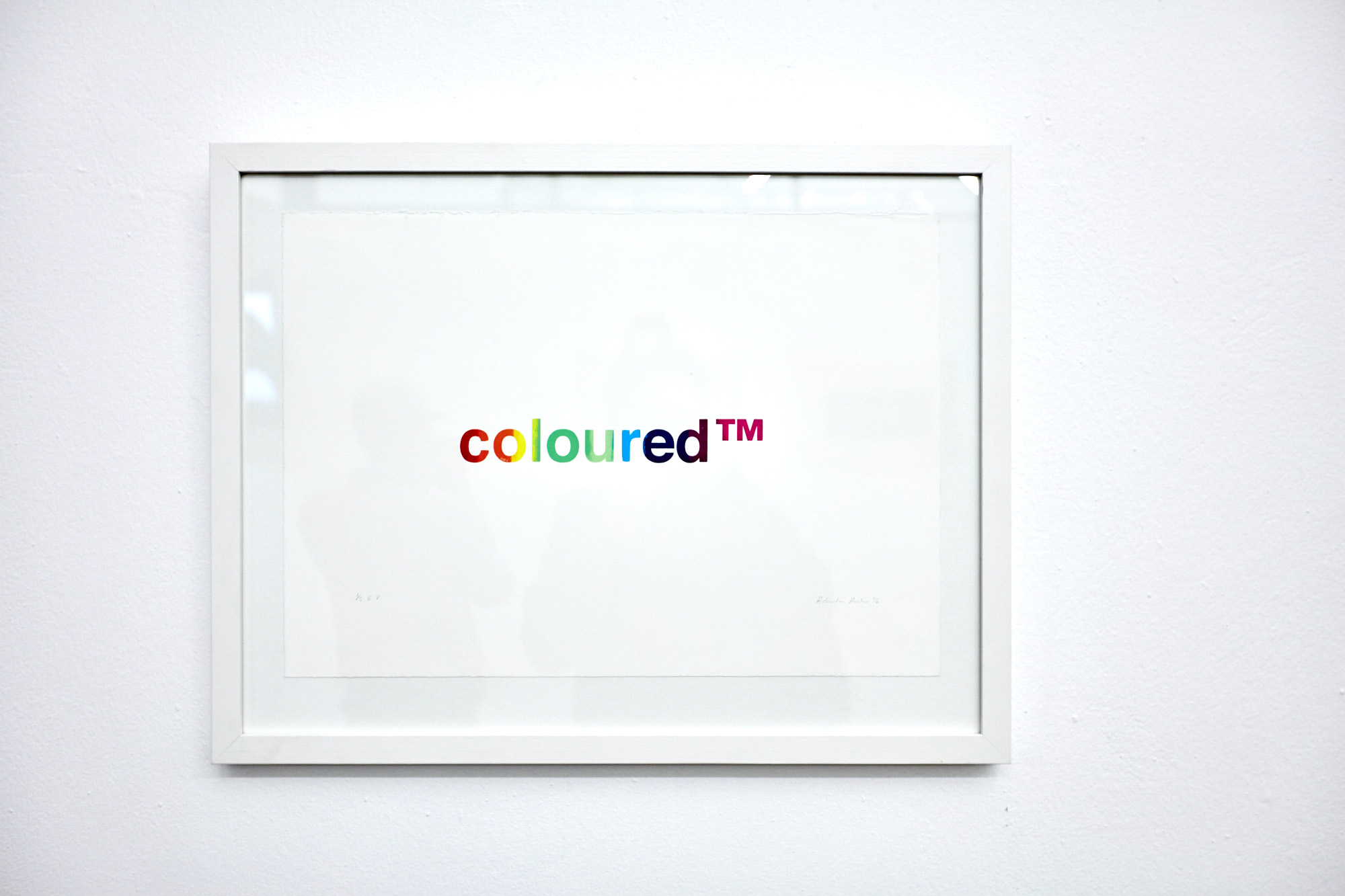 A white-framed print on a white wall showing the single word "coloured™" in rainbow-gradient lowercase letters centred on white paper, with the artist's pencil signature below.