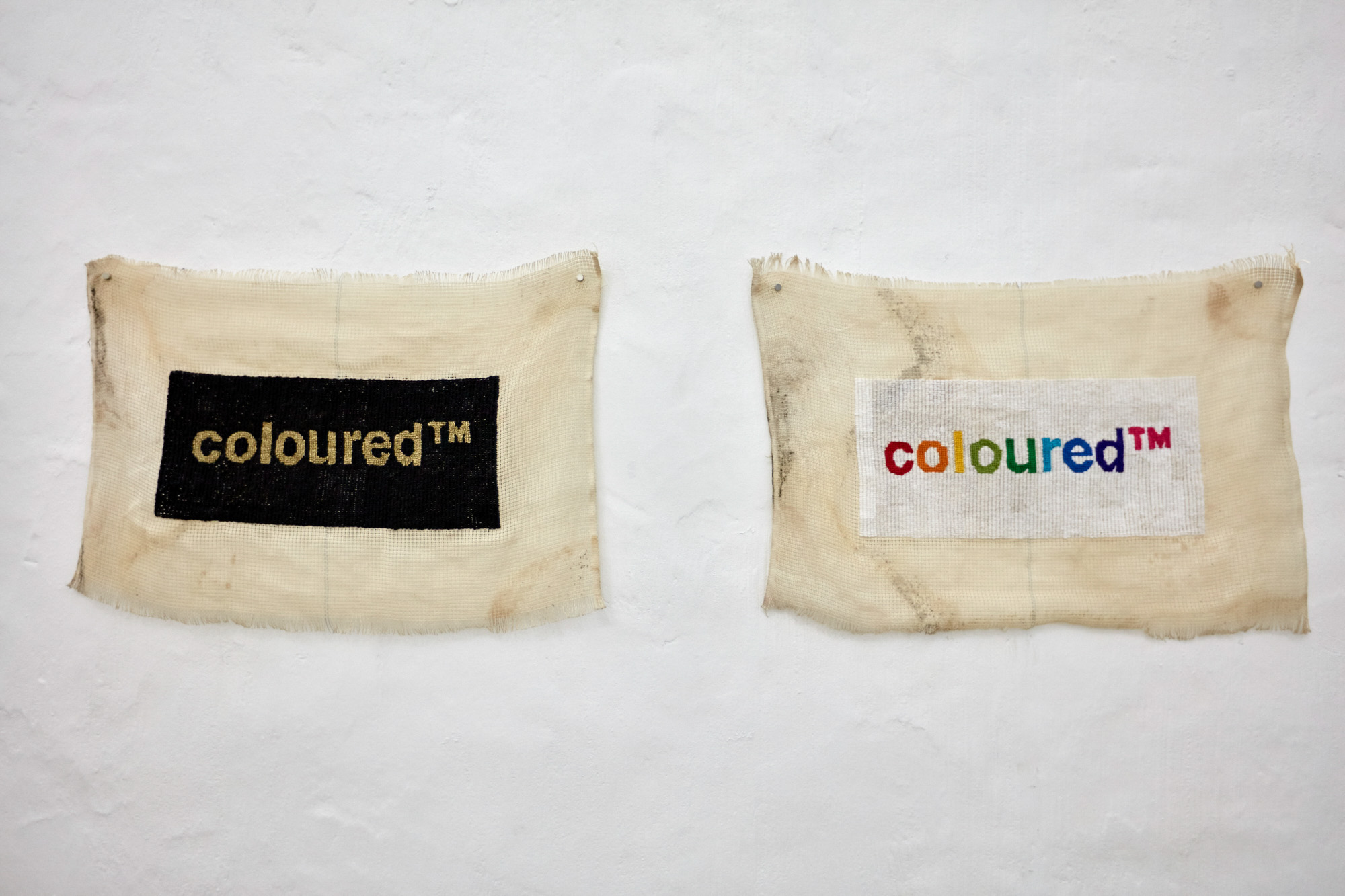 Two frayed, tea-stained, needlepoint canvas tapestries pinned to a white brick wall. The left shows "coloured™" in gold thread on a black rectangle; the right shows "coloured™" stitched in rainbow letters on white cotton.