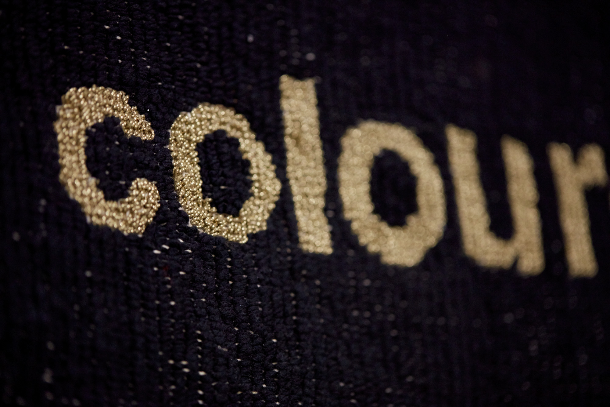 Macro detail of a tapestry showing part of the word "coloured" stitched in shimmering metallic gold thread against dense black wool.