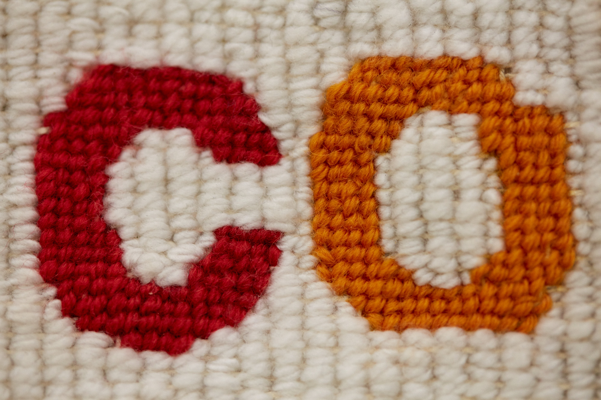 Macro detail of needlepoint stitching showing the letters "c" and "o" in red and orange wool on a cream woven background.