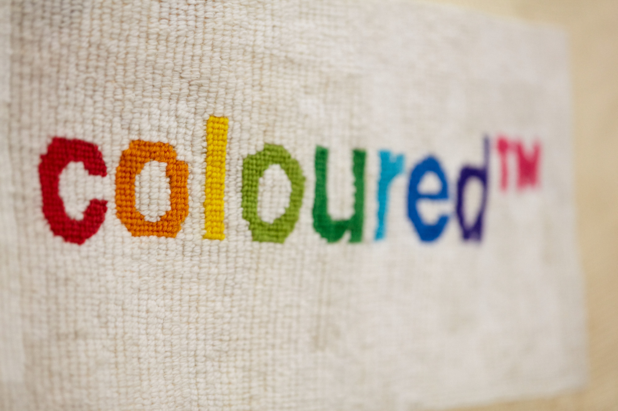 Close-up of the word "coloured™" stitched in rainbow-coloured thread — red, orange, yellow, green, blue, purple, and pink — surrounded by white needlepoint cotton on needlepoint canvas.