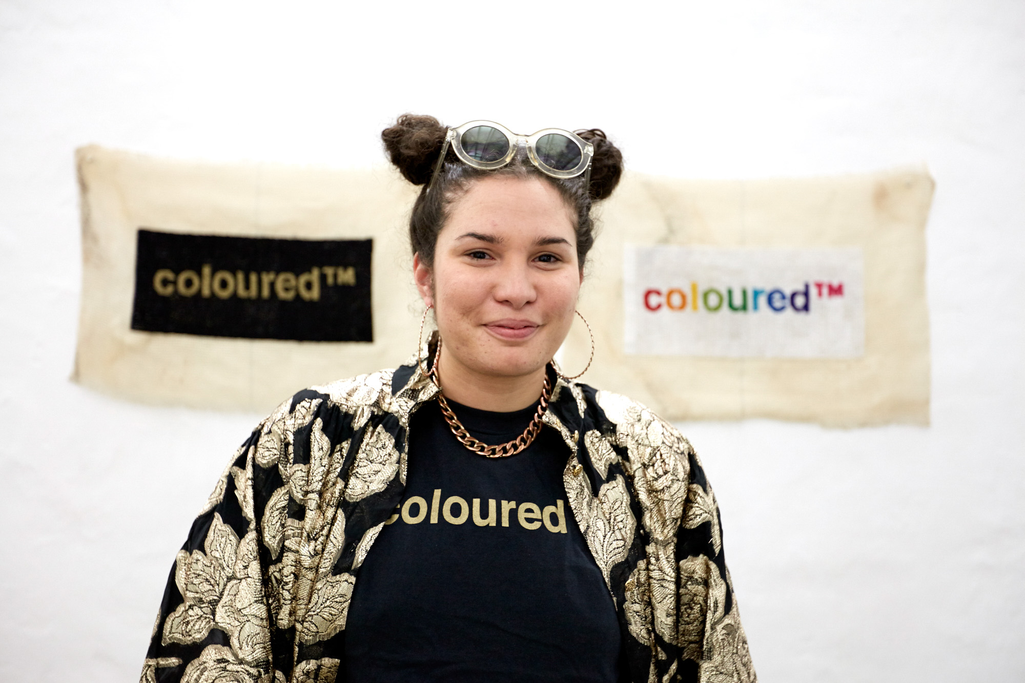 Roberta Joy Rich smiles at the camera wearing a black "coloured" T-shirt, gold brocade jacket, hoop earrings, and sunglasses atop her hair, standing before her two "coloured™" tapestries.