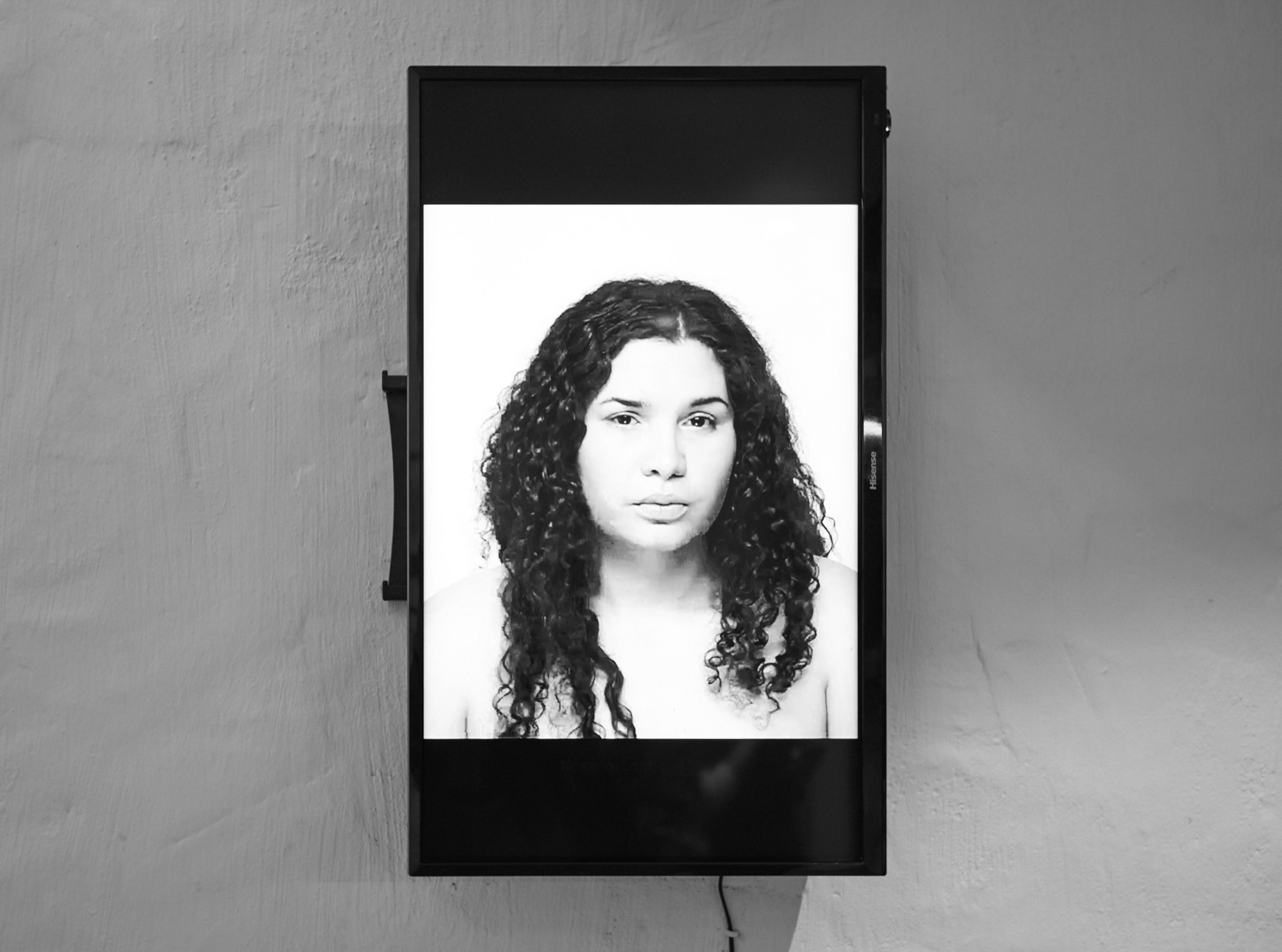 A wall-mounted screen displays a black-and-white frontal portrait of a woman - the artist Roberta Joy Rich - with long curly hair gazing at the camera, is part of a video work in the exhibition.