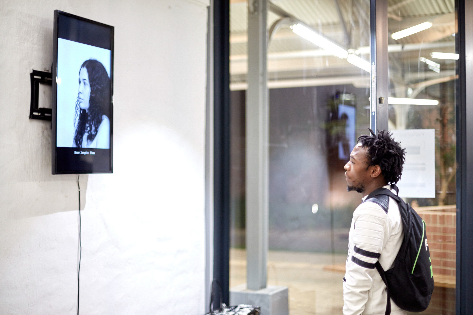 An African male visitor with a backpack watches a wall-mounted video showing a black-and-white profile portrait of a woman, with a caption reading "Brow length: 55mm" beneath the image.