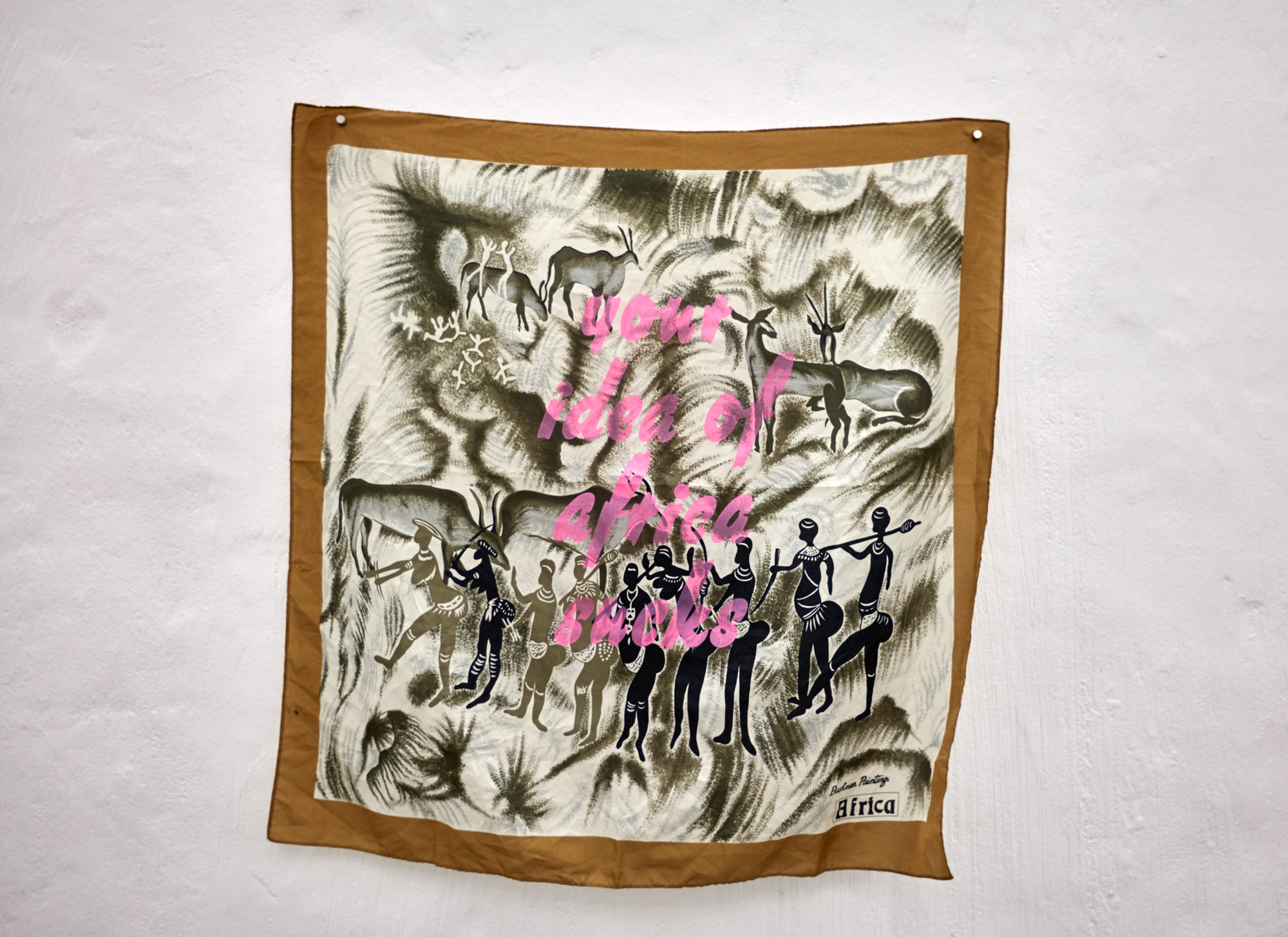 A vintage silk souvenir scarf pinned to a white wall, printed with African animals and Indigenous figures in olive tones, with "Bushman Paintings Africa" in one corner. The words "your idea of africa sucks" are overprinted in pink cursive.