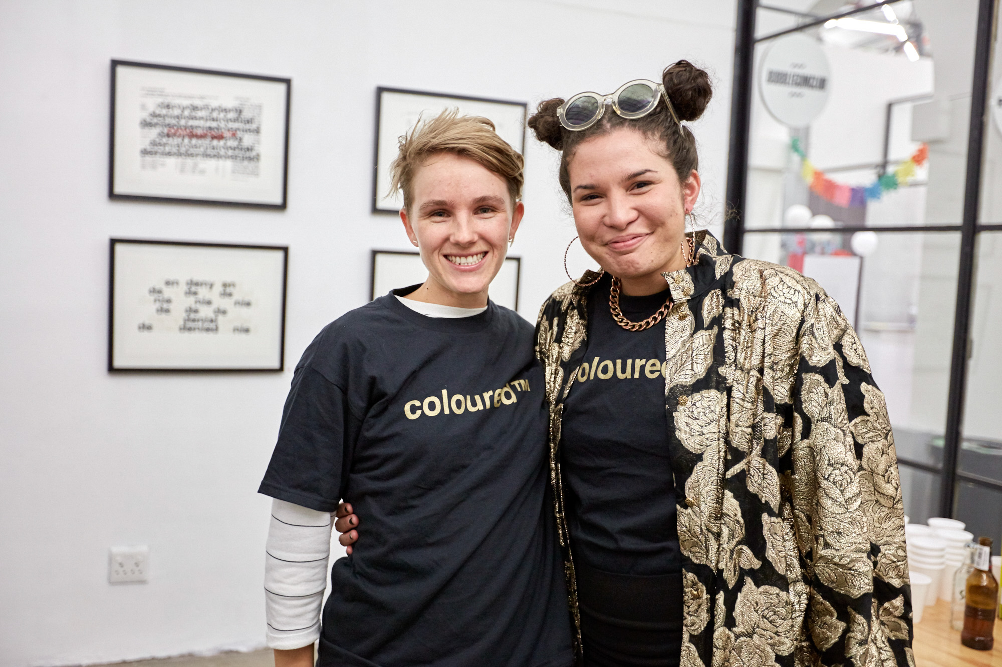 Two smiling artists pose arm in arm wearing black "coloured™" t-shirts with gold lettering. Behind them hang framed prints repeating the words "deny, denial, denied".