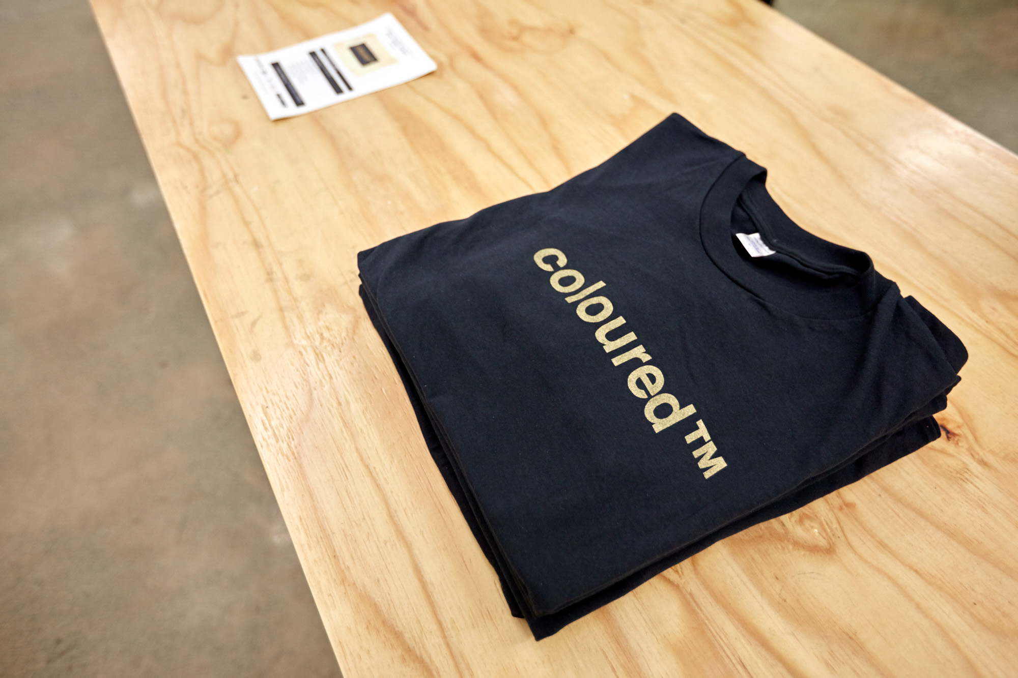 A folded black t-shirt printed with "coloured™" in gold lettering lies on a wooden table beside an exhibition pamphlet.