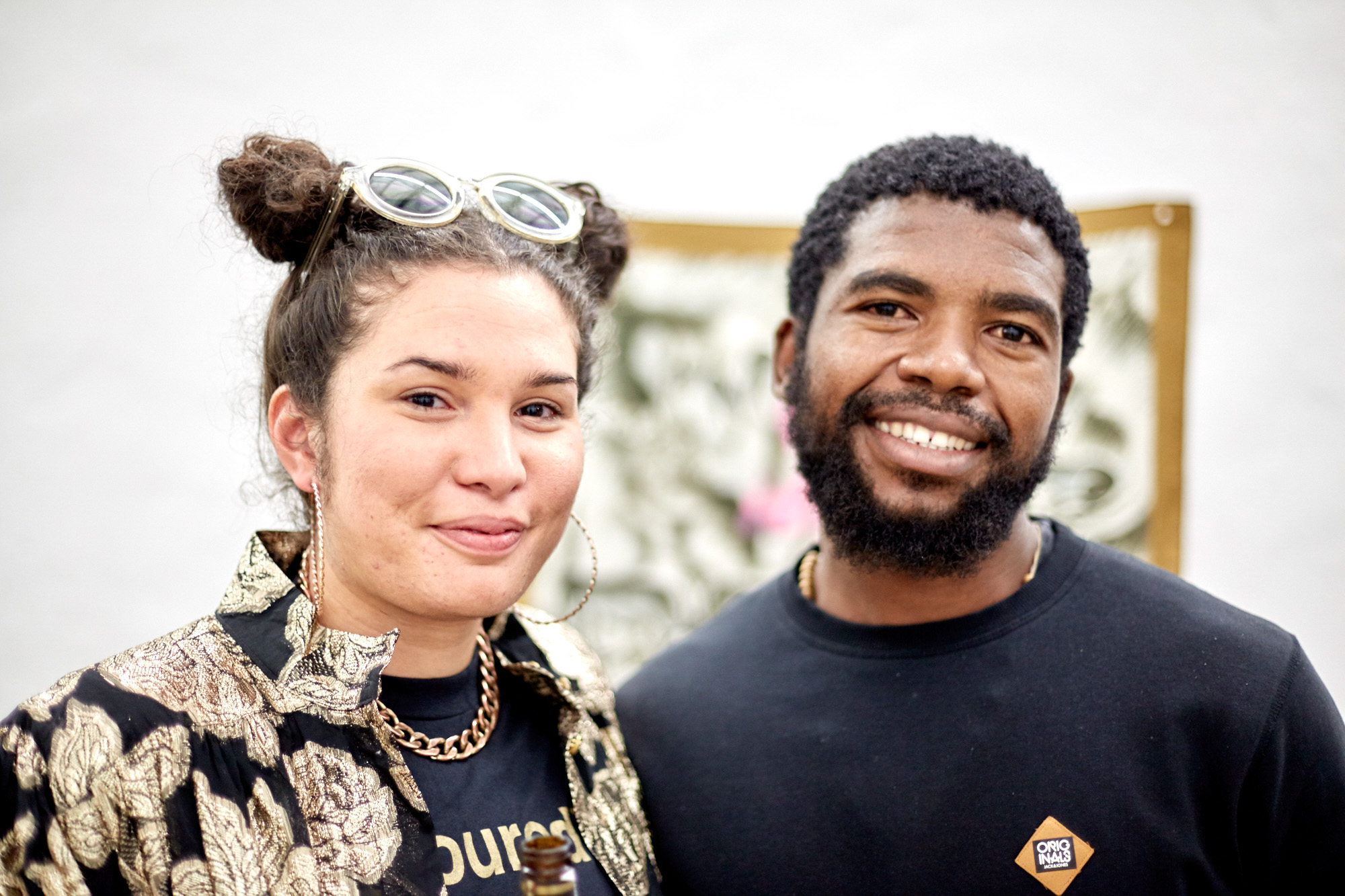 A woman - artist Roberta Joy Rich - wearing space buns, hoop earrings and a gold brocade jacket stands beside a smiling bearded African man in a black sweater, with the scarf artwork blurred behind them.