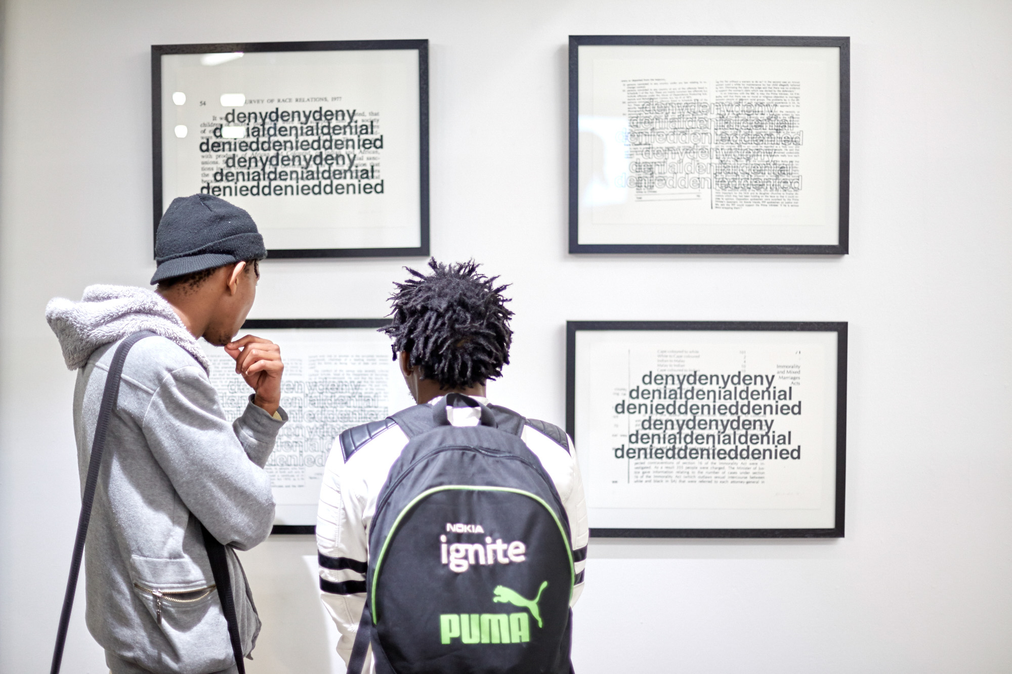 Two young African men study four black-framed prints in which the words "deny, denial, denied" are printed in bold type over pages from a 1977 survey of race relations.