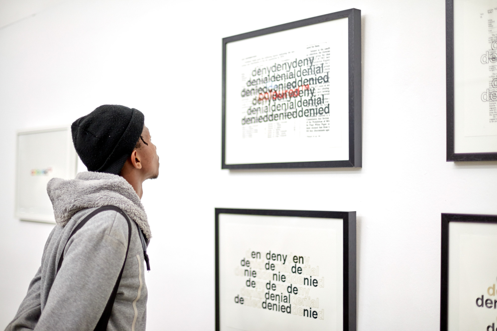 A young African man in a black beanie and grey sweatshirt looks up at a framed print where "coloured™" appears in red within layers of repeated "deny, denial, denied" black text.
