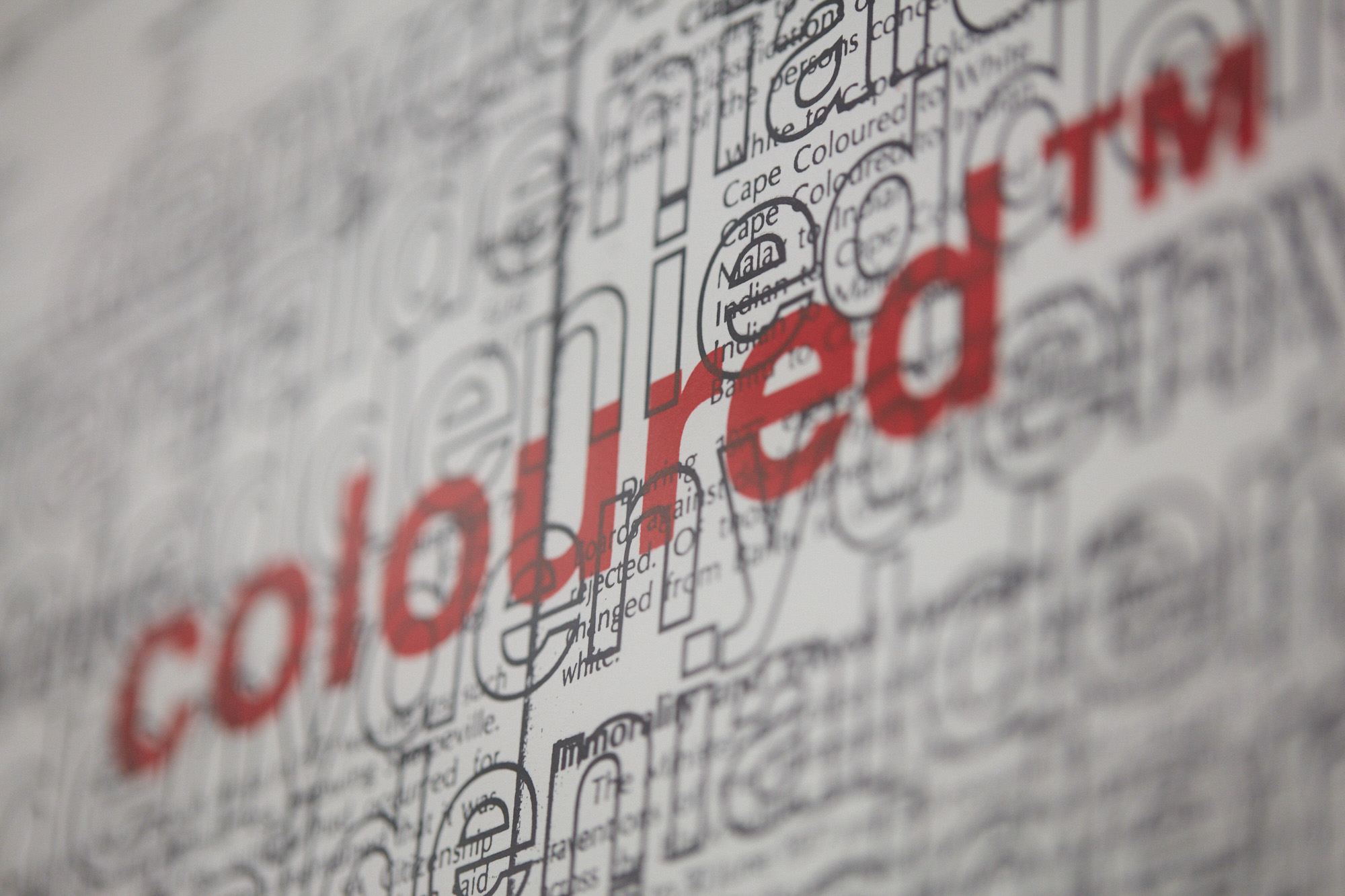 Close-up of a print: "coloured™" in red overlaps dense layers of black type listing apartheid-era racial reclassifications such as "Cape Coloured to White".