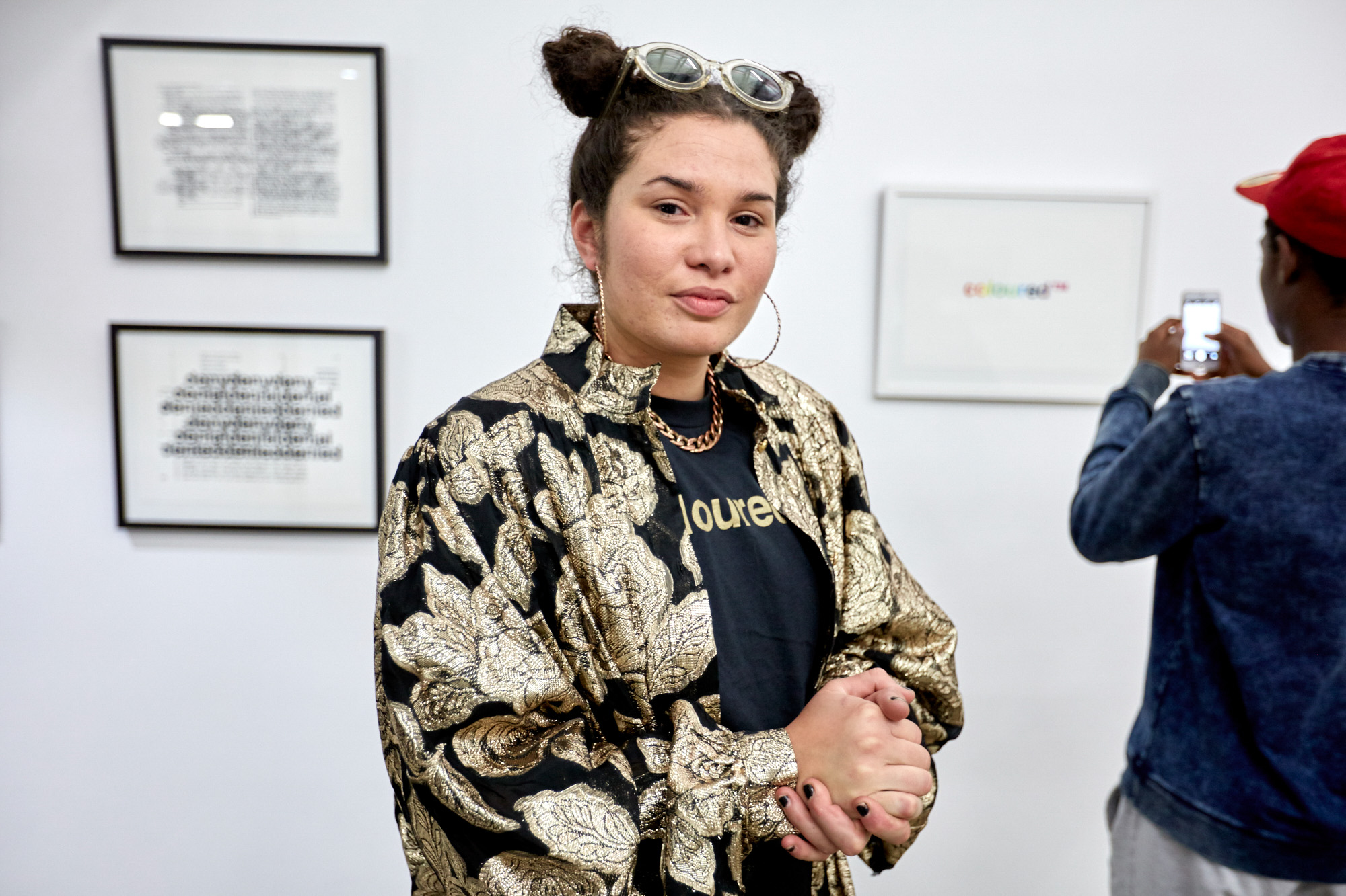 The artist in a gold brocade jacket and "coloured™" t-shirt clasps her hands and looks at the camera; behind her a visitor photographs the rainbow "coloured™" print.