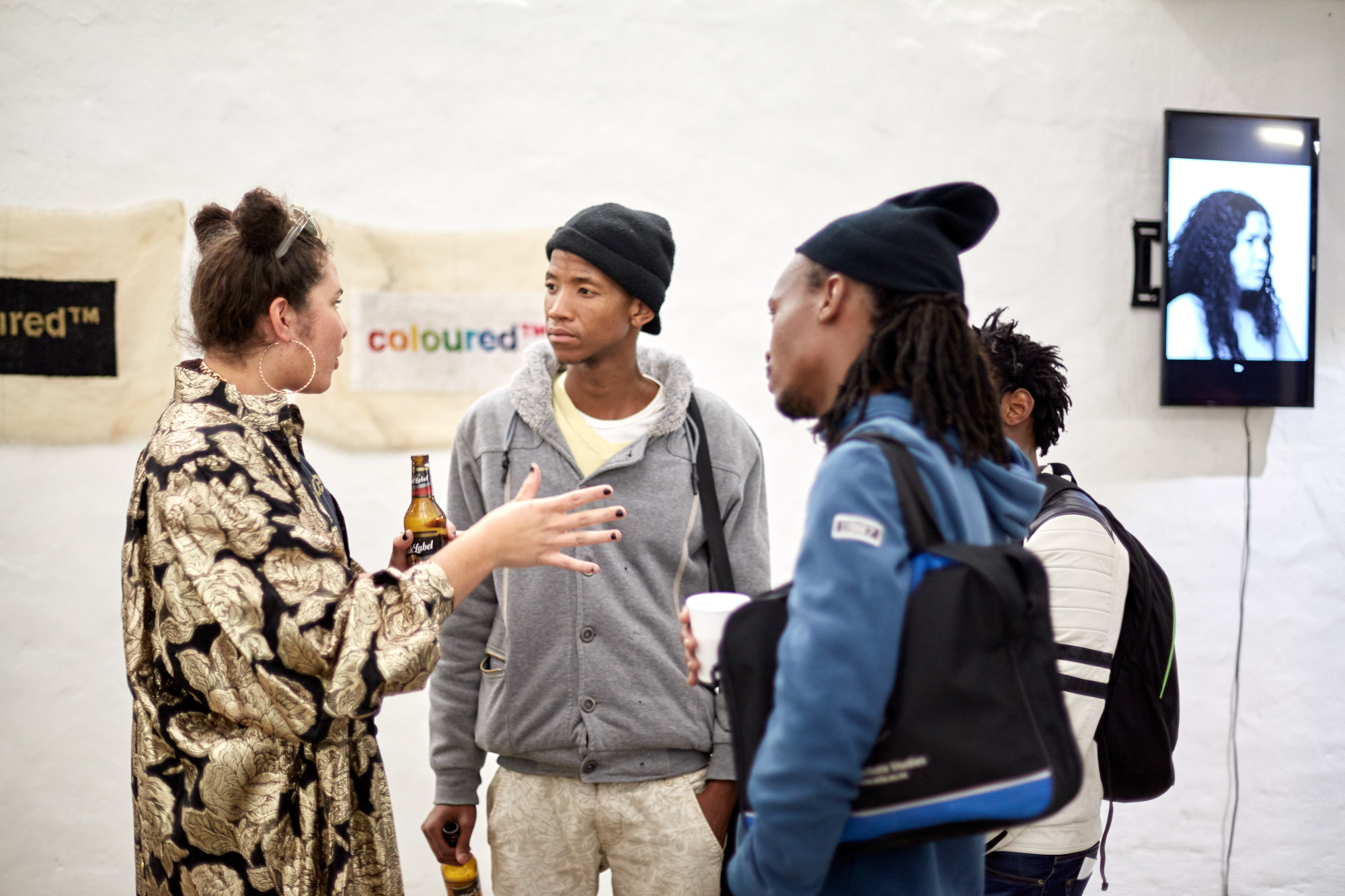 The artist gestures while talking with three African male visitors wearing beanies and sweatshirts. Behind them hang "coloured™" tapestries and a screen showing a black-and-white portrait of a woman- the artist in her video work.