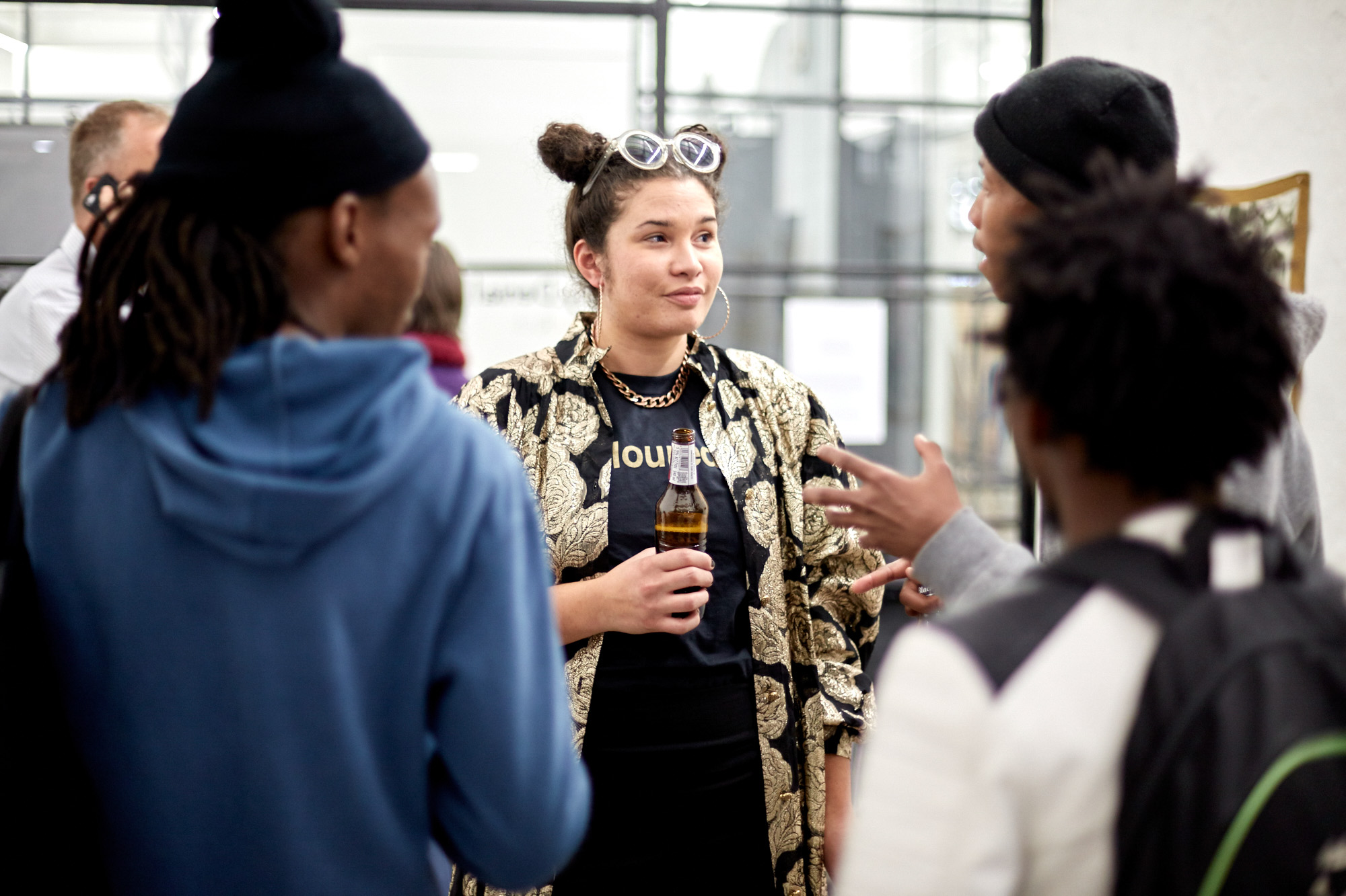 The artist, holding a Black Label bottle, chats with a group of African male visitors wearing beanies and hoodies inside the bright gallery.