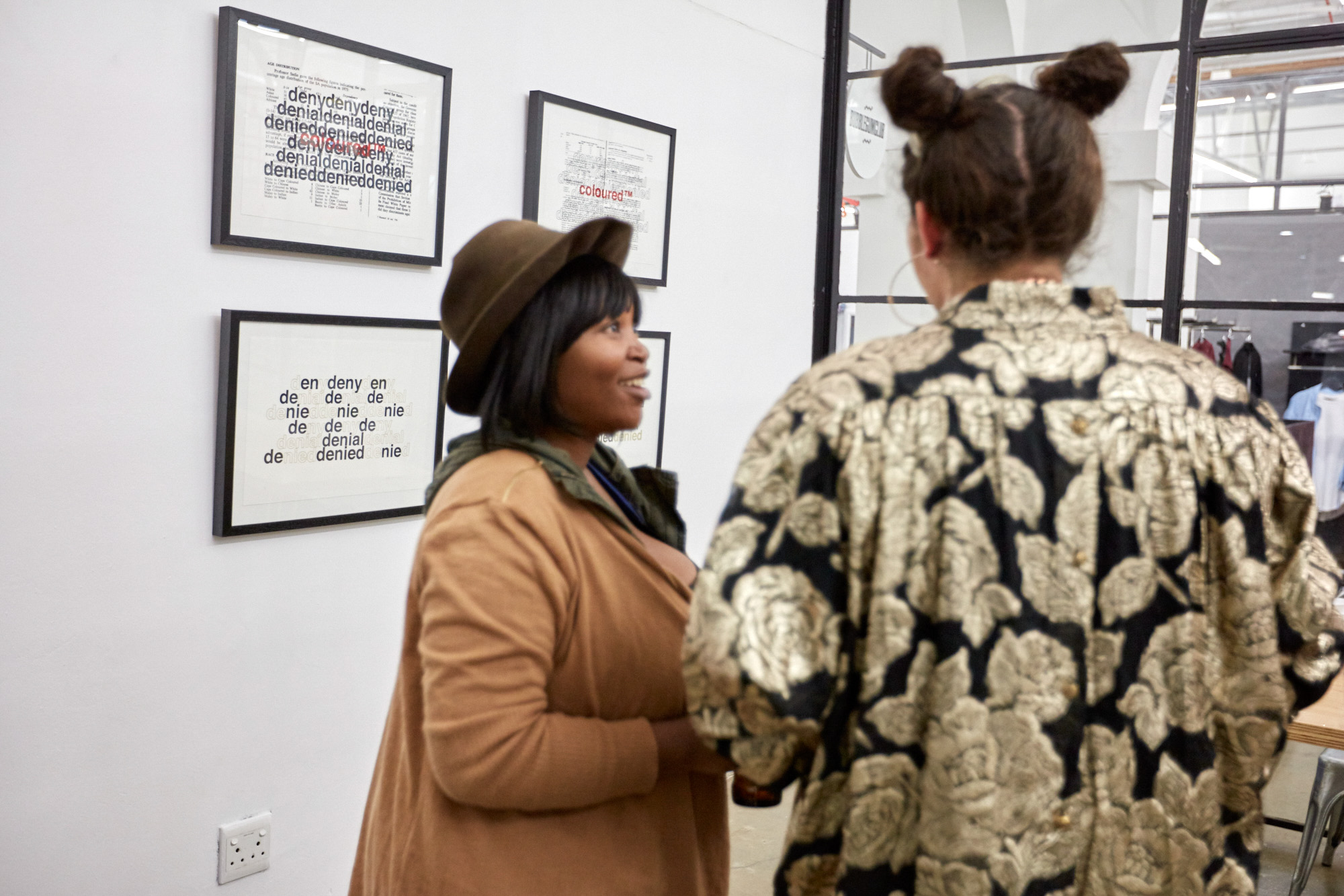 Two African women talk in front of black-framed "deny, denial, denied" prints; one wears a brown hat and coat, the other a gold rose-patterned brocade jacket.