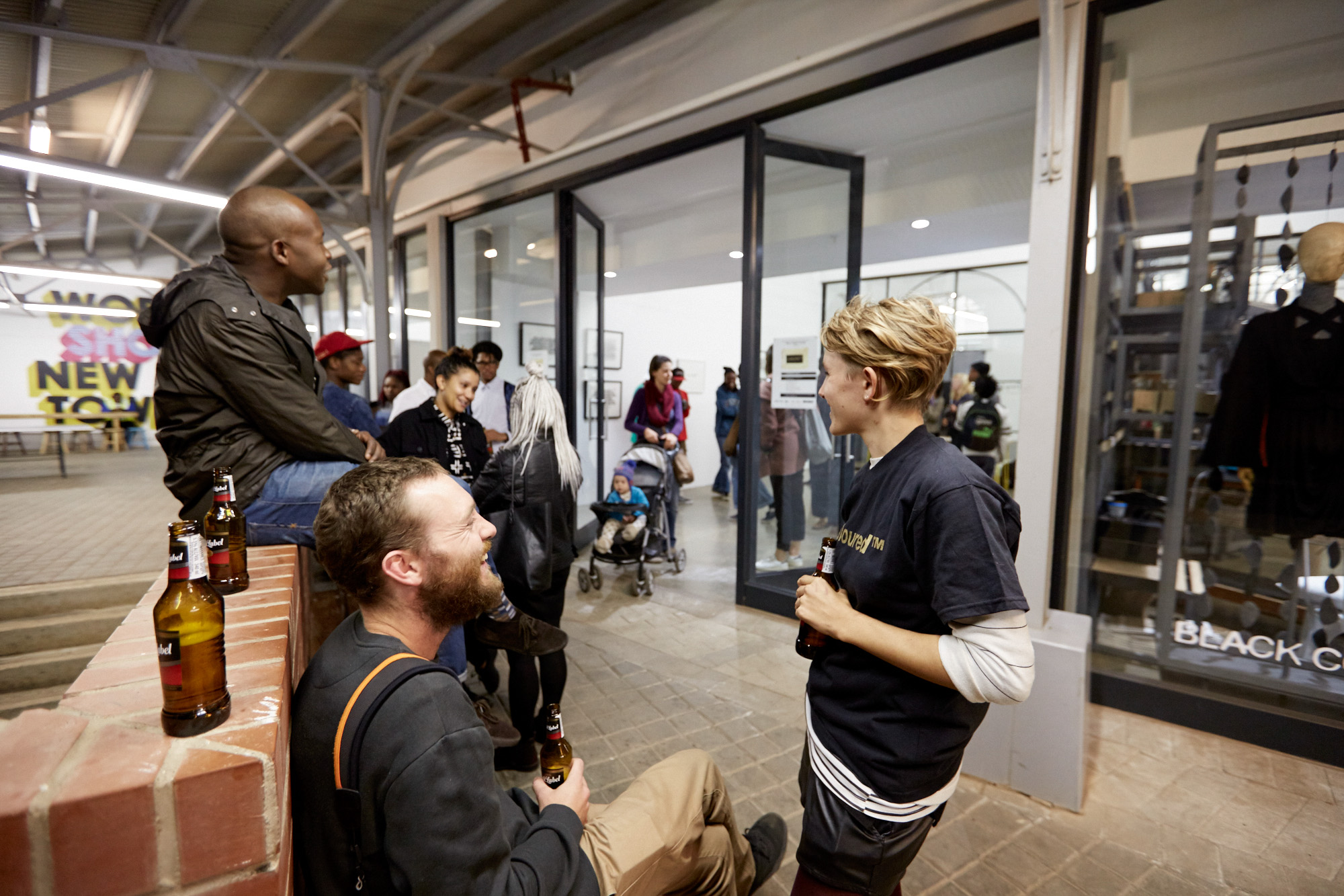 Visitors of various ethnicities socialise with drinks outside the glass-fronted gallery, some seated and standing upon the brick paving; a mural reading "Workshop Newtown" is visible in the background.
