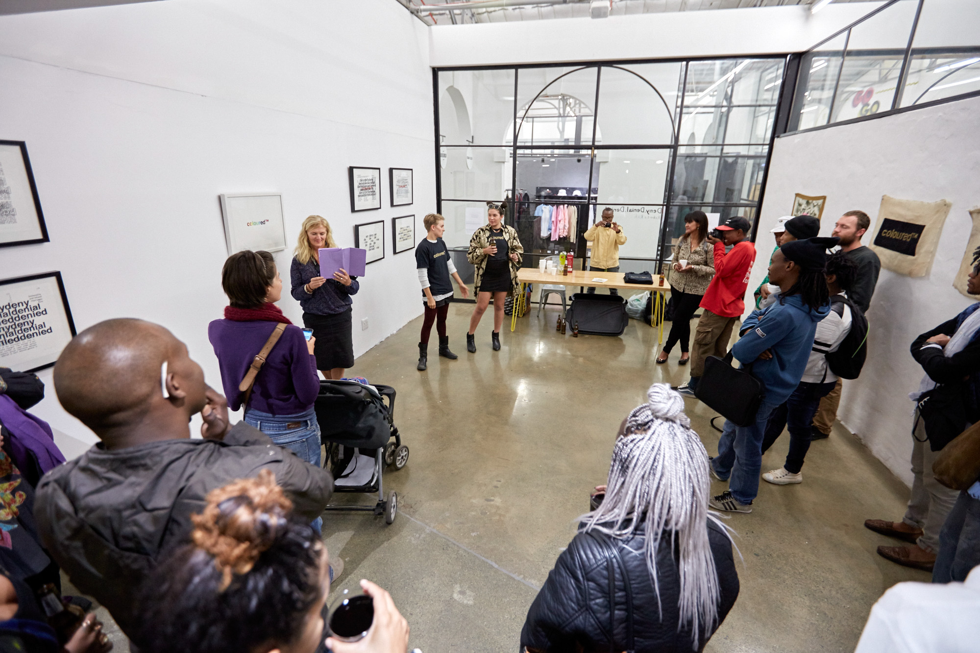 A crowd circles the gallery listening to two female artists wearing black tshirts that read "coloured™" in gold, address the room. A woman is taking photos from a purple tablet stands beside the two artists during the exhibition opening.