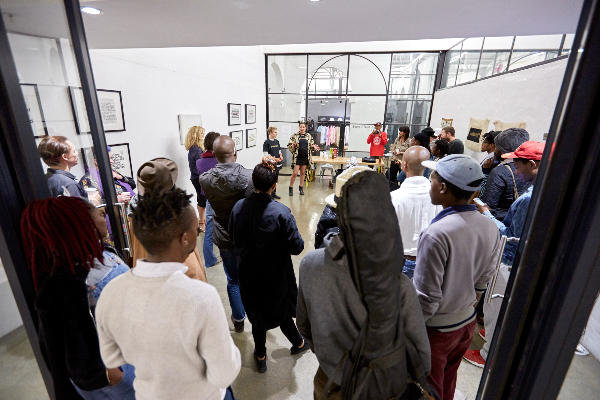 View through glass doors of a packed gallery exhibition opening: dozens of visitors face the artists, who address the crowd beside a wood table. An African man in a red jumper stands behind the table while holding a camera towards the crowd.
