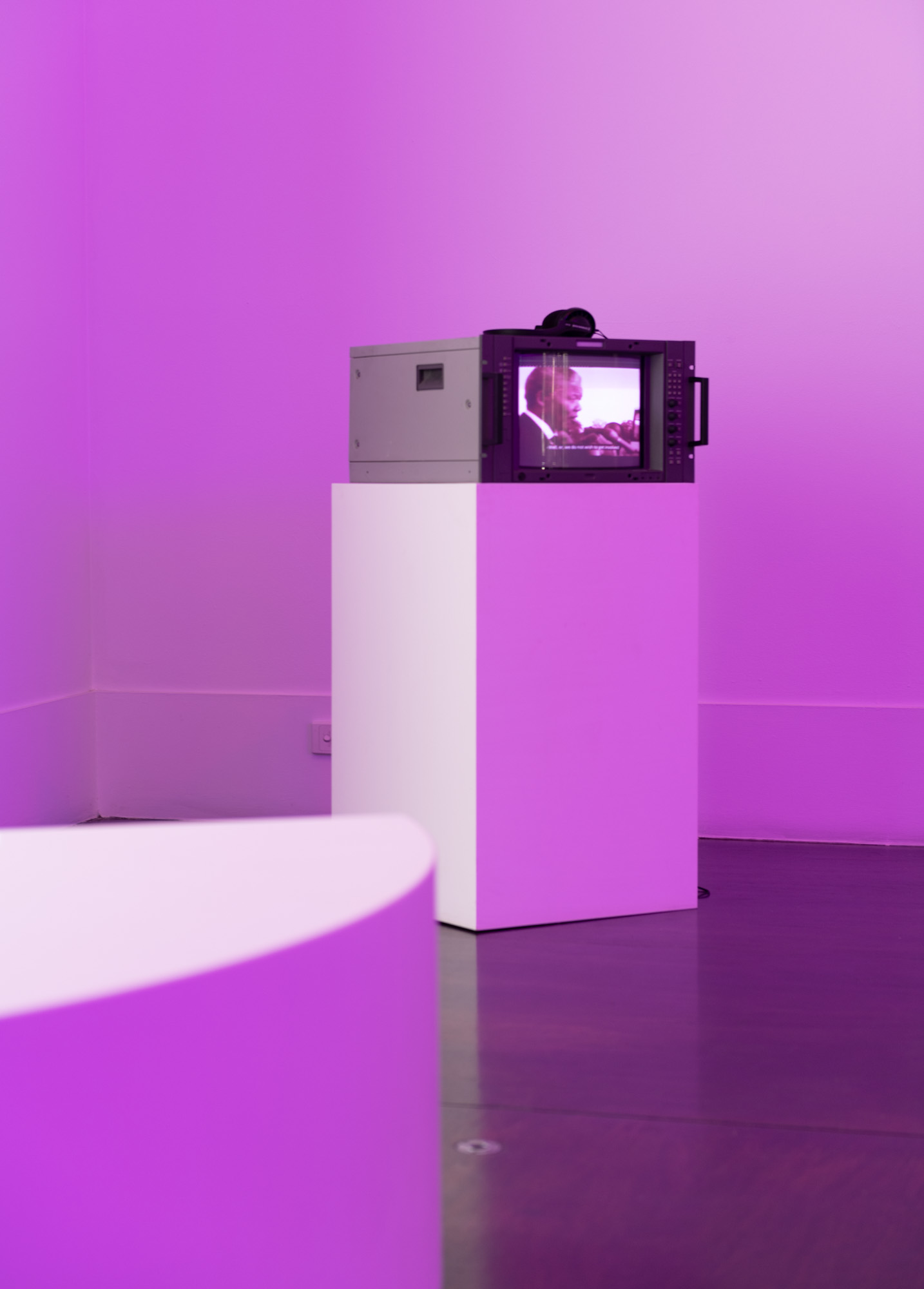 Viewed past a curved white bench, a broadcast monitor with headphones sits on a white plinth in the corner of a gallery bathed in magenta light, playing subtitled archival footage of Nelson Mandela speaking into microphones.