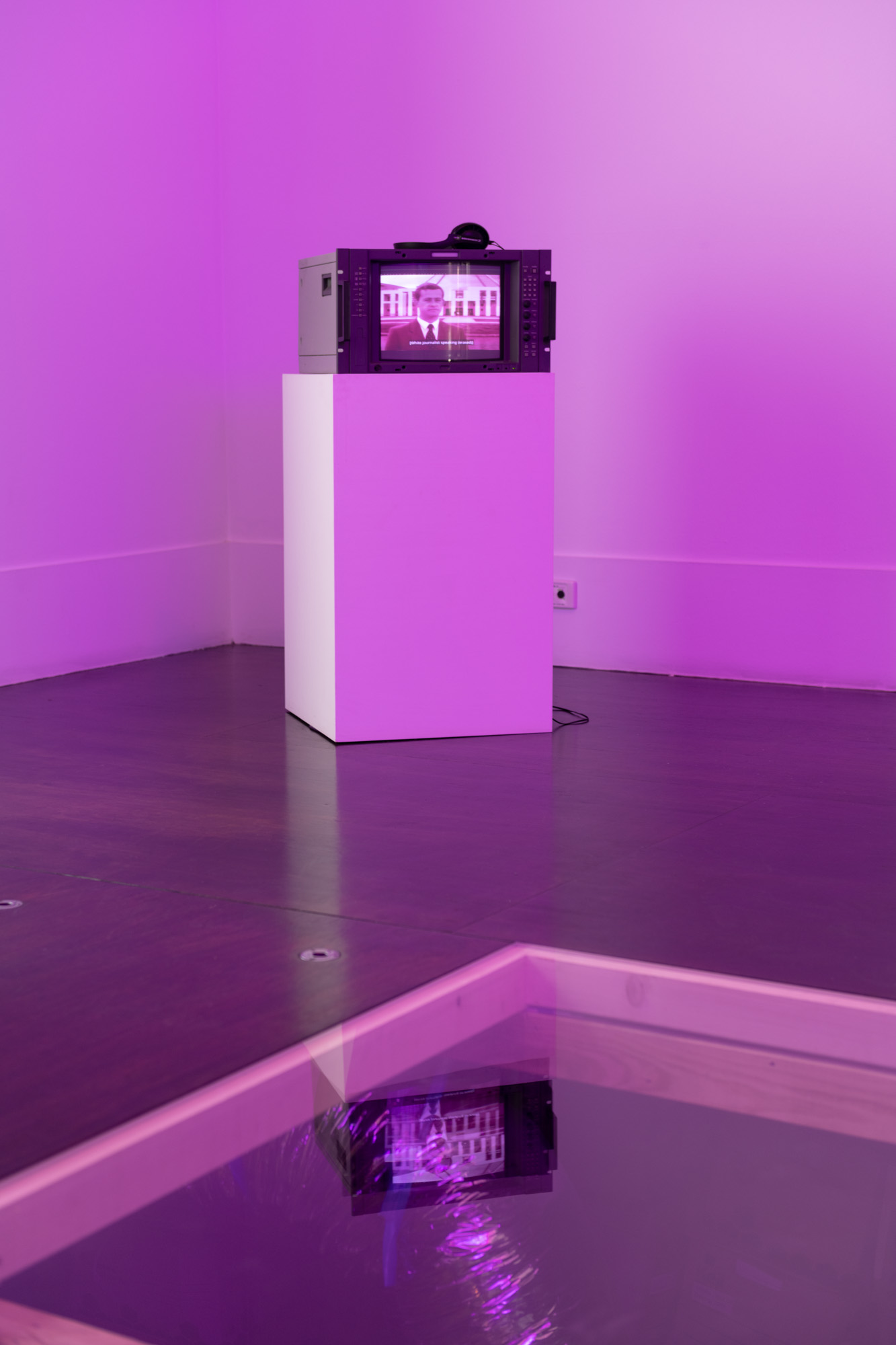 In a gallery bathed in pink-purple light, a broadcast monitor with headphones sits on a white plinth playing subtitled footage of a journalist outside Australian Parliament House. The screen is reflected upon transparent perspex set in the floor in the foreground.