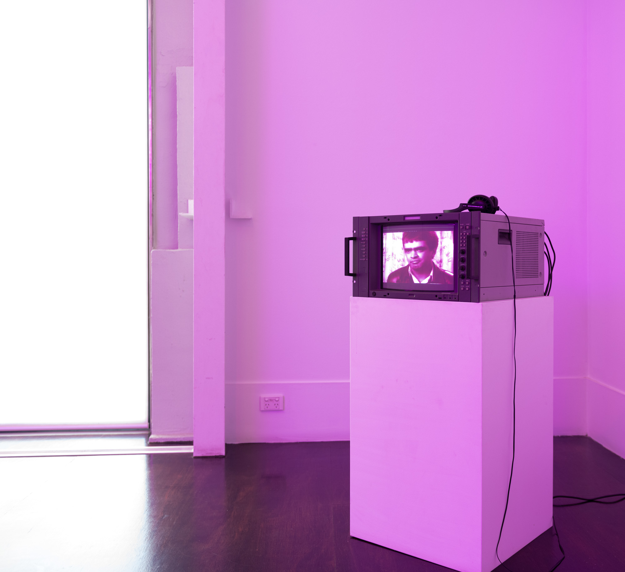 A vintage broadcast monitor topped with headphones sits on a white plinth in a corner bathed in magenta light. On screen, a subtitled video shows activist Dr. Gary Foley, a man with dark hair wearing a leather jacket in front of patterned curtains.