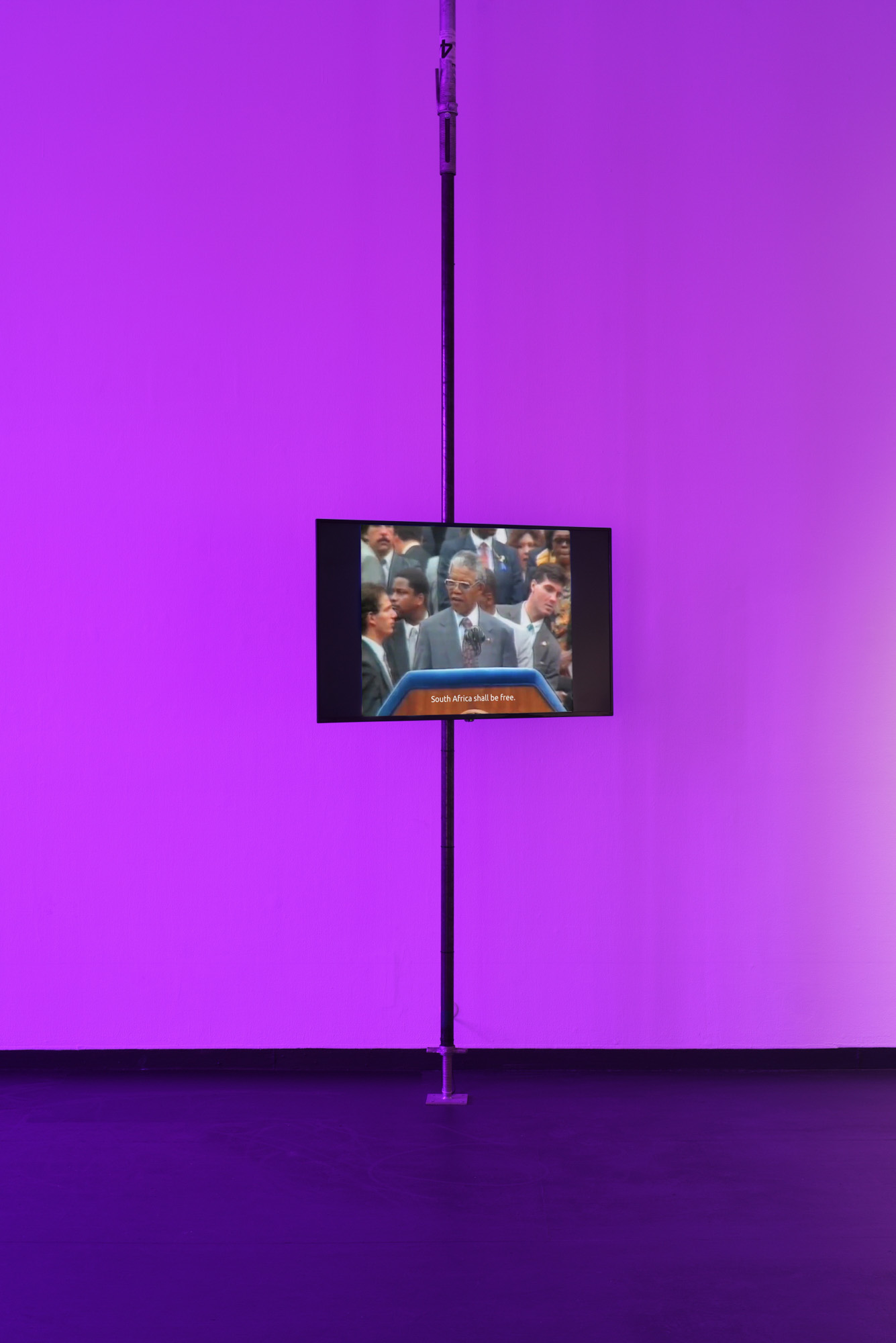 A flat-screen mounted on a floor-to-ceiling pole in a violet-lit space plays archival footage of Nelson Mandela at a podium, subtitled "South Africa shall be free."