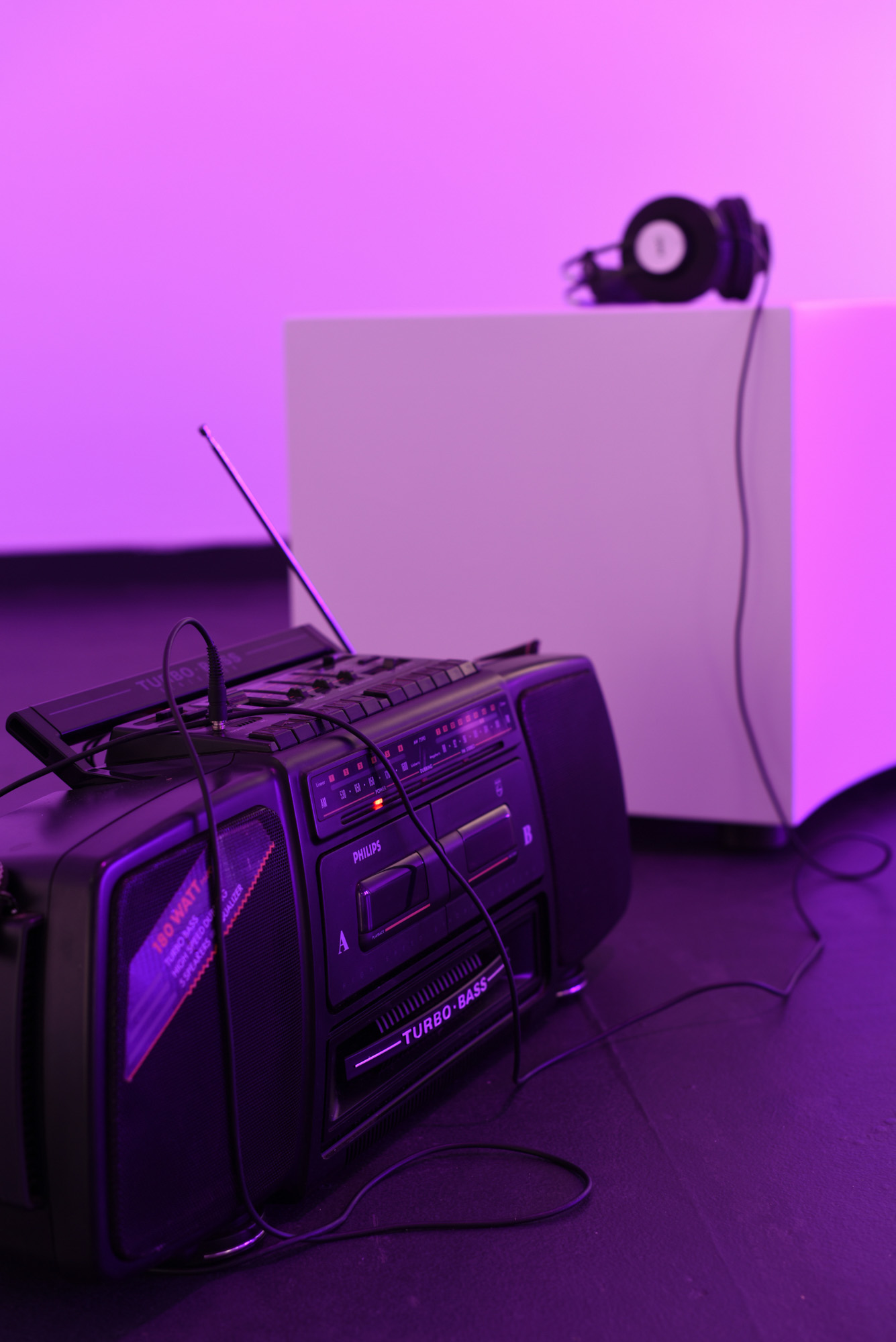 A black Philips "Turbo Bass" radio boombox with twin cassette decks and an extended aerial sits on the floor of a violet-lit gallery, headphone cable plugged in and draped over it. Behind it, a pair of black headphones rests on top of a white plinth- also bathed in purple light- and headphone cord hanging down it's side.