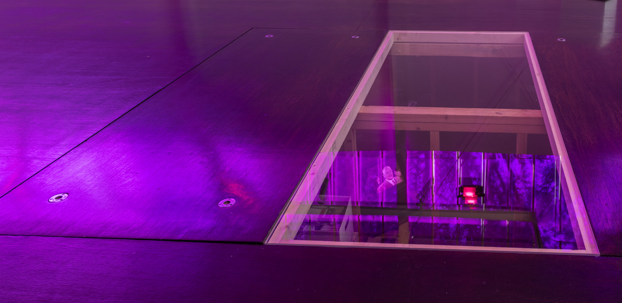 A rectangular glass panel set into the dark timber gallery floor, glowing under purple light. Through the glass, timber framing and a sub-floor space are visible, with a small broadcast monitor showing a man- Hendrik Verwoerd- in a suit within reflections of windows and a red-filtered light fixture upon the glass' surface.