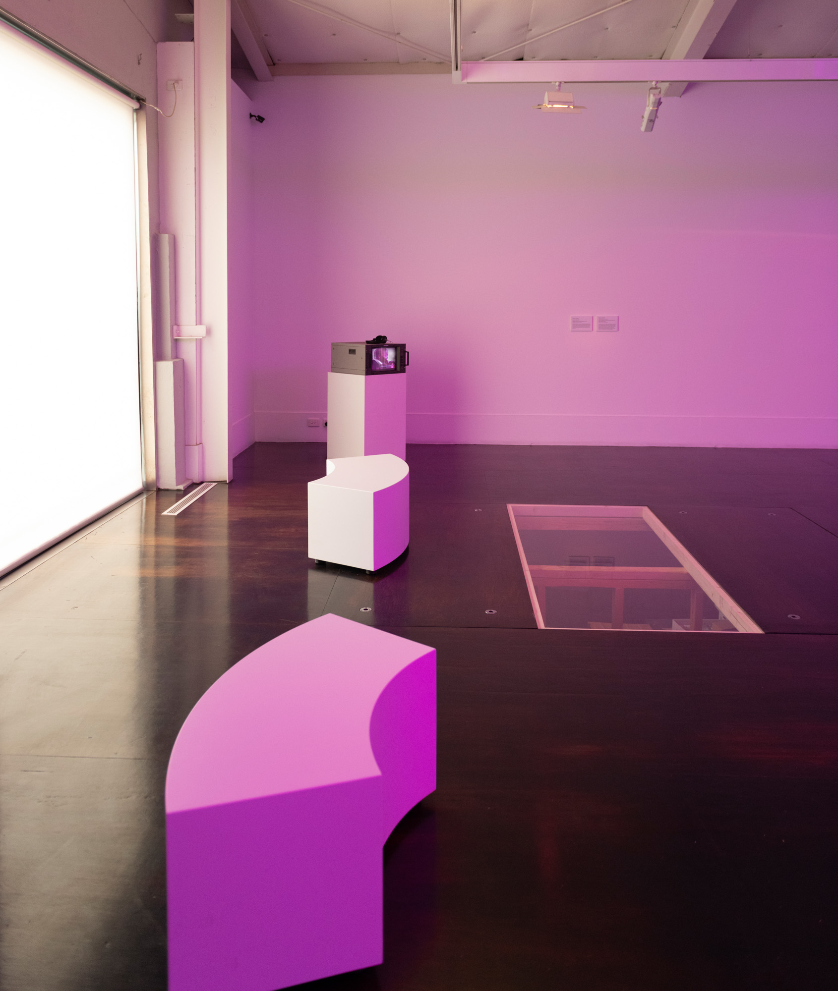 Wide view of a purple-lit gallery: a broadcast monitor topped with headphones sits on a white plinth in the corner, faced by two curved white benches, while a glass viewing panel is set flush into the dark wood floor at right. Daylight glows through a screened window at left.