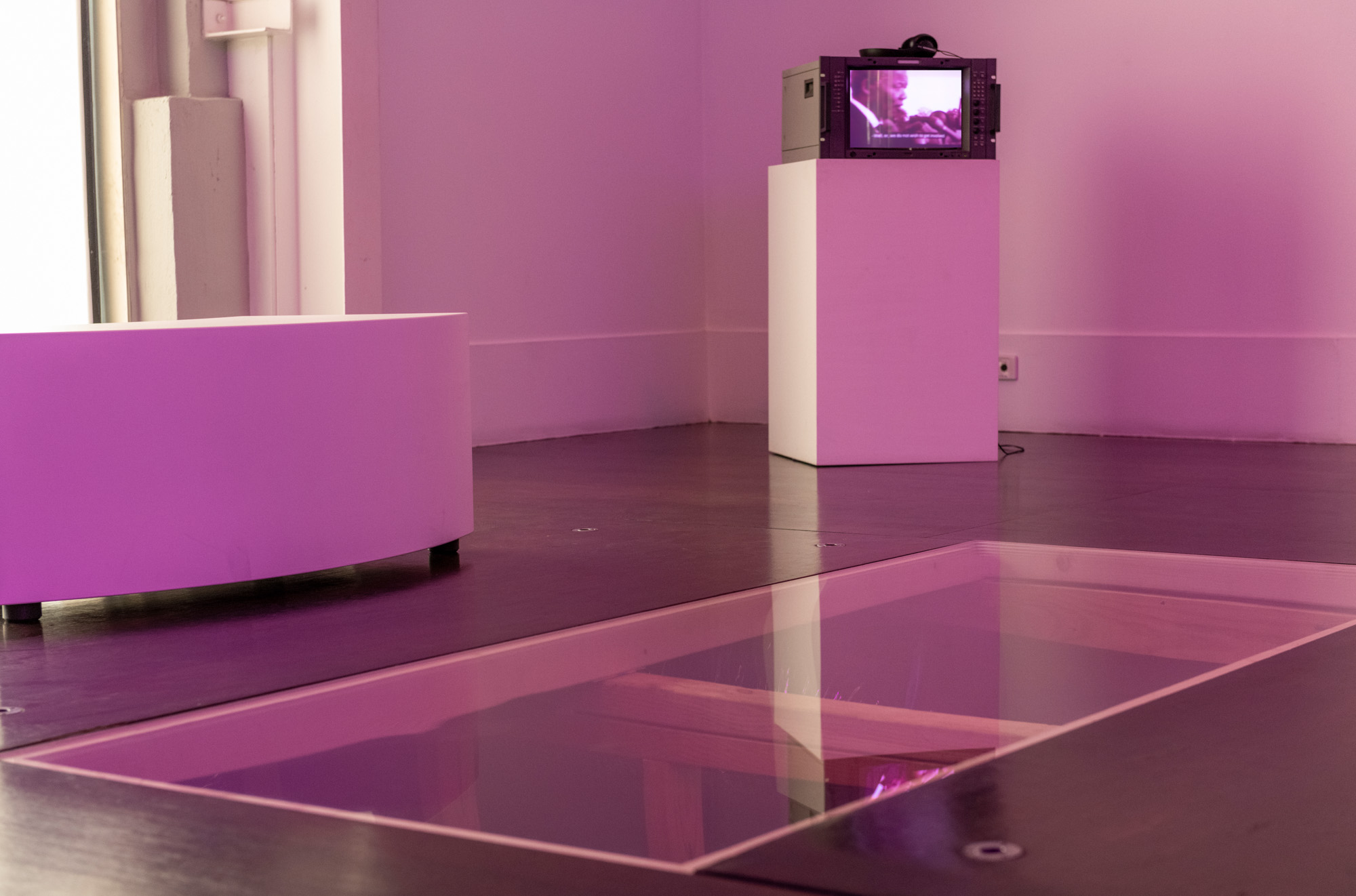 Low view across the glass floor panel toward the corner plinth, where a broadcast monitor plays subtitled footage of Nelson Mandela speaking into a bank of microphones. Timber joists are faintly visible beneath the tinted glass in the foreground, with hints of purple light illuminating the space.