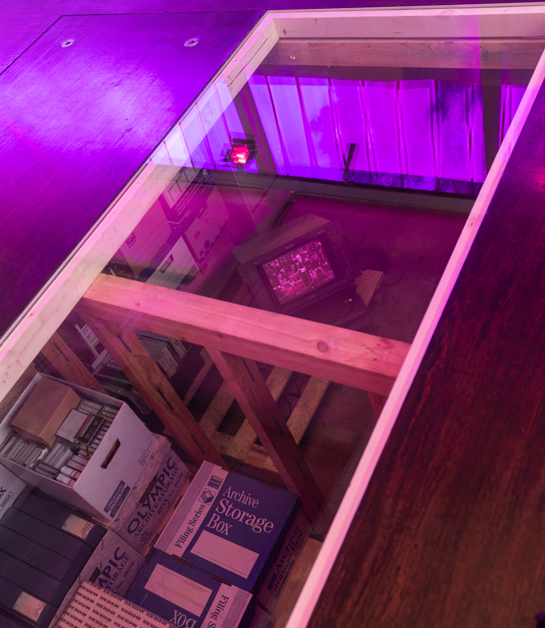 Looking directly down through the glass floor panel into an under-floor archive: timber framing, stacked archive storage boxes and cases of cassette tapes surround a Sony broadcast monitor playing black-and-white footage of a crowd, all washed in pink and purple light.