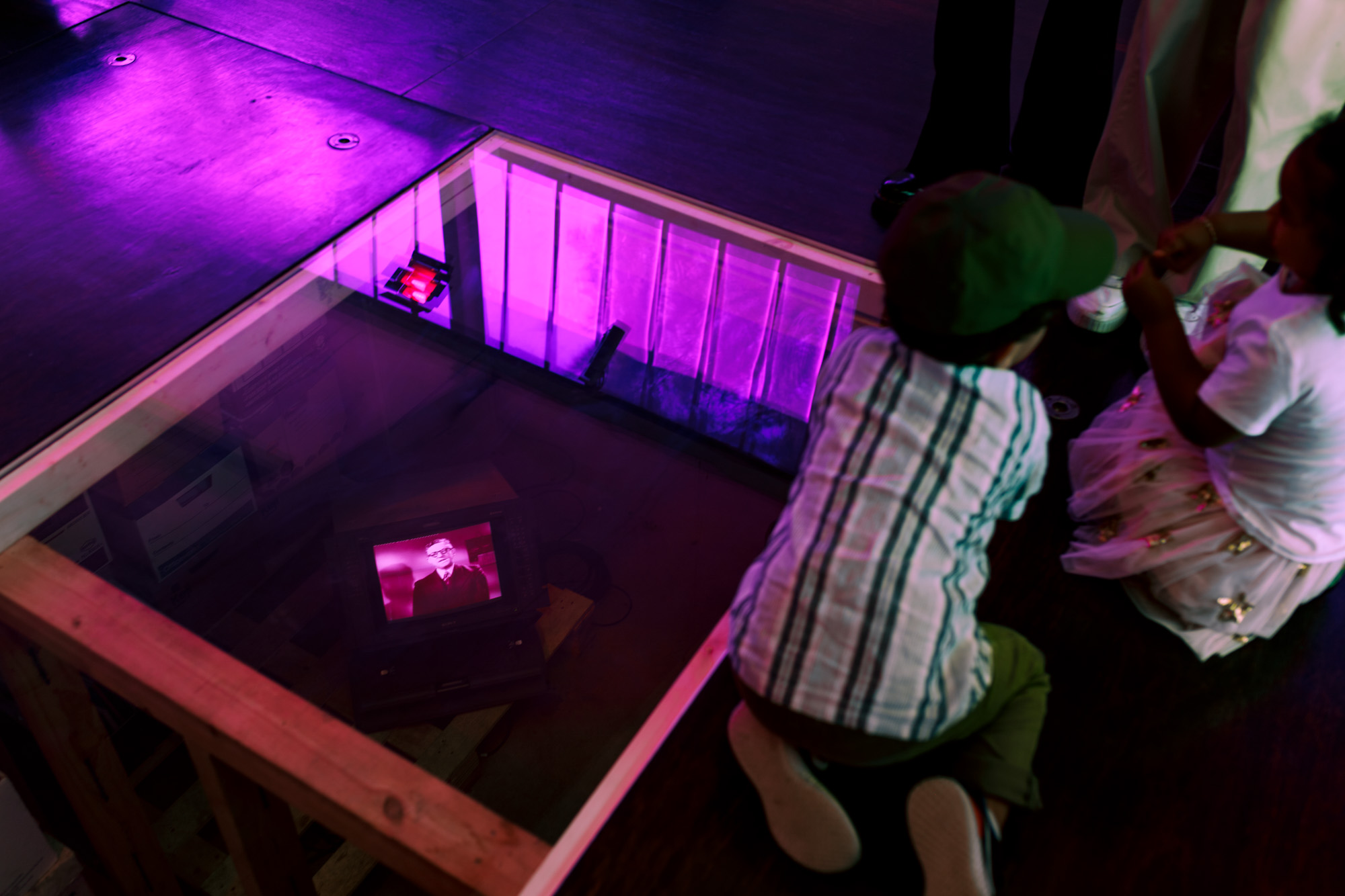 Two children kneel at the edge of the glass floor panel, peering down at the Sony broadcast monitor below, which shows a bespectacled man in a suit- Arthur Calwell. Reflected pink window light fills the glass around them.
