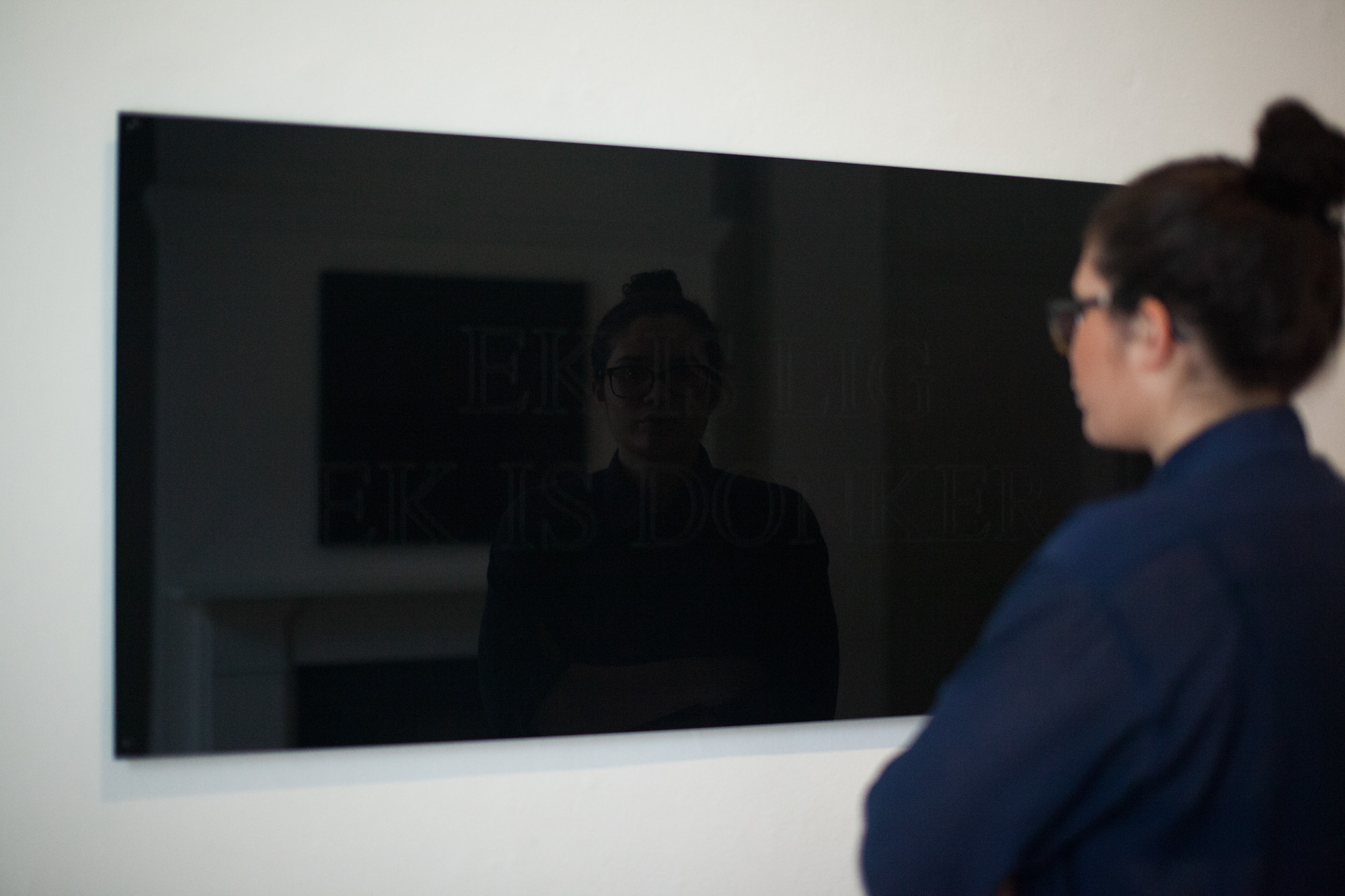 A viewer with dark brown hair tied up, wearing a navy collar shirt with folded arms looks into a black mirrored text work, their face appearing in its dark reflection.