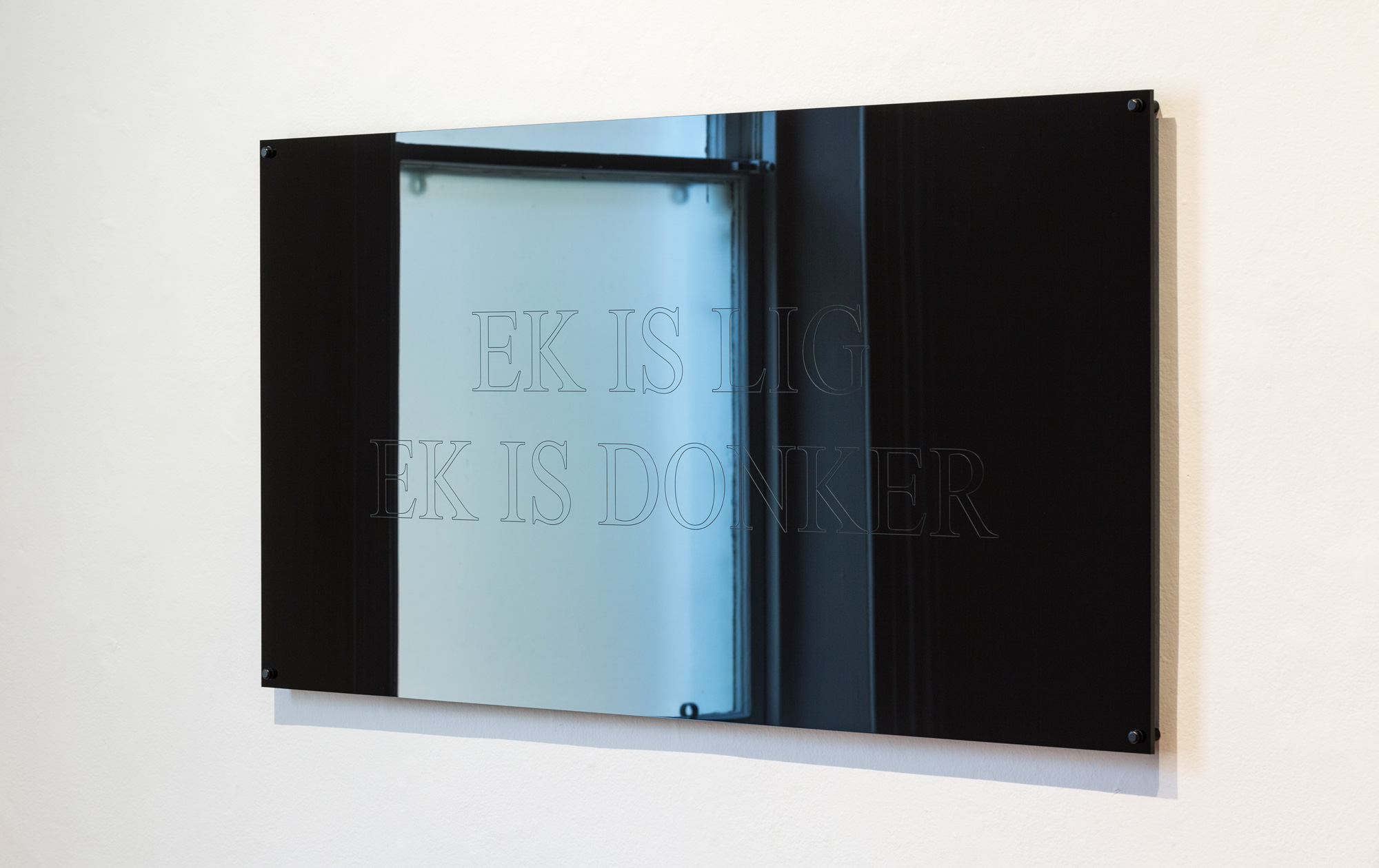 Black mirrored panel engraved with the Afrikaans text “EK IS LIG / EK IS DONKER,” reflecting a blue-lit window.