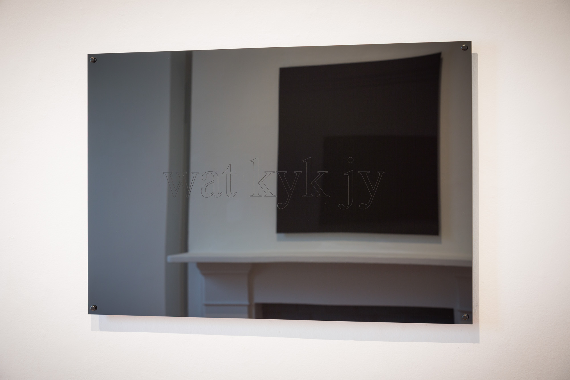 Black mirrored panel engraved “wat kyk jy,” reflecting a fireplace and another dark artwork.