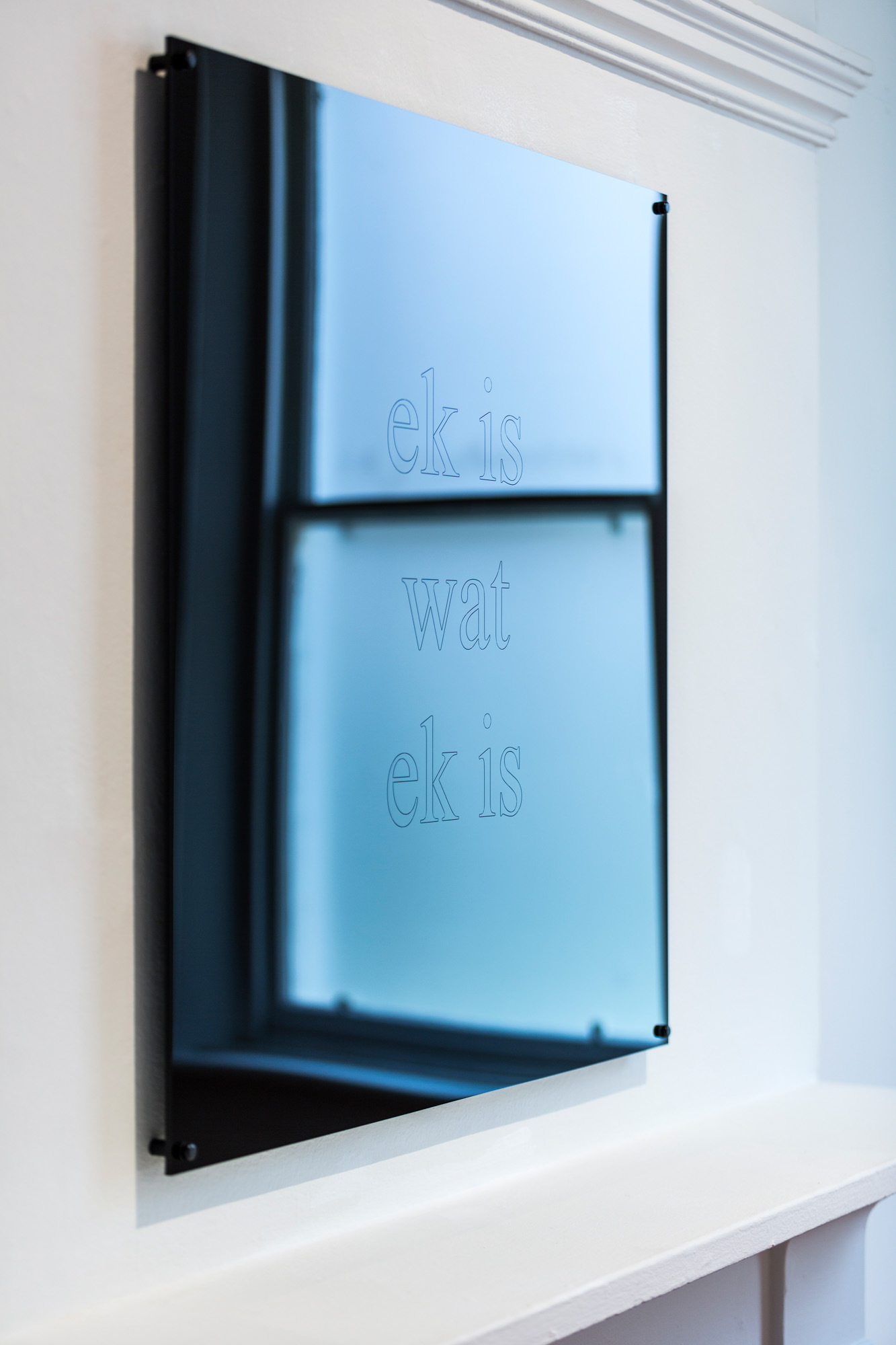 Angled detail of the mirrored work “ek is / wat / ek is,” reflecting a window in shades of blue.