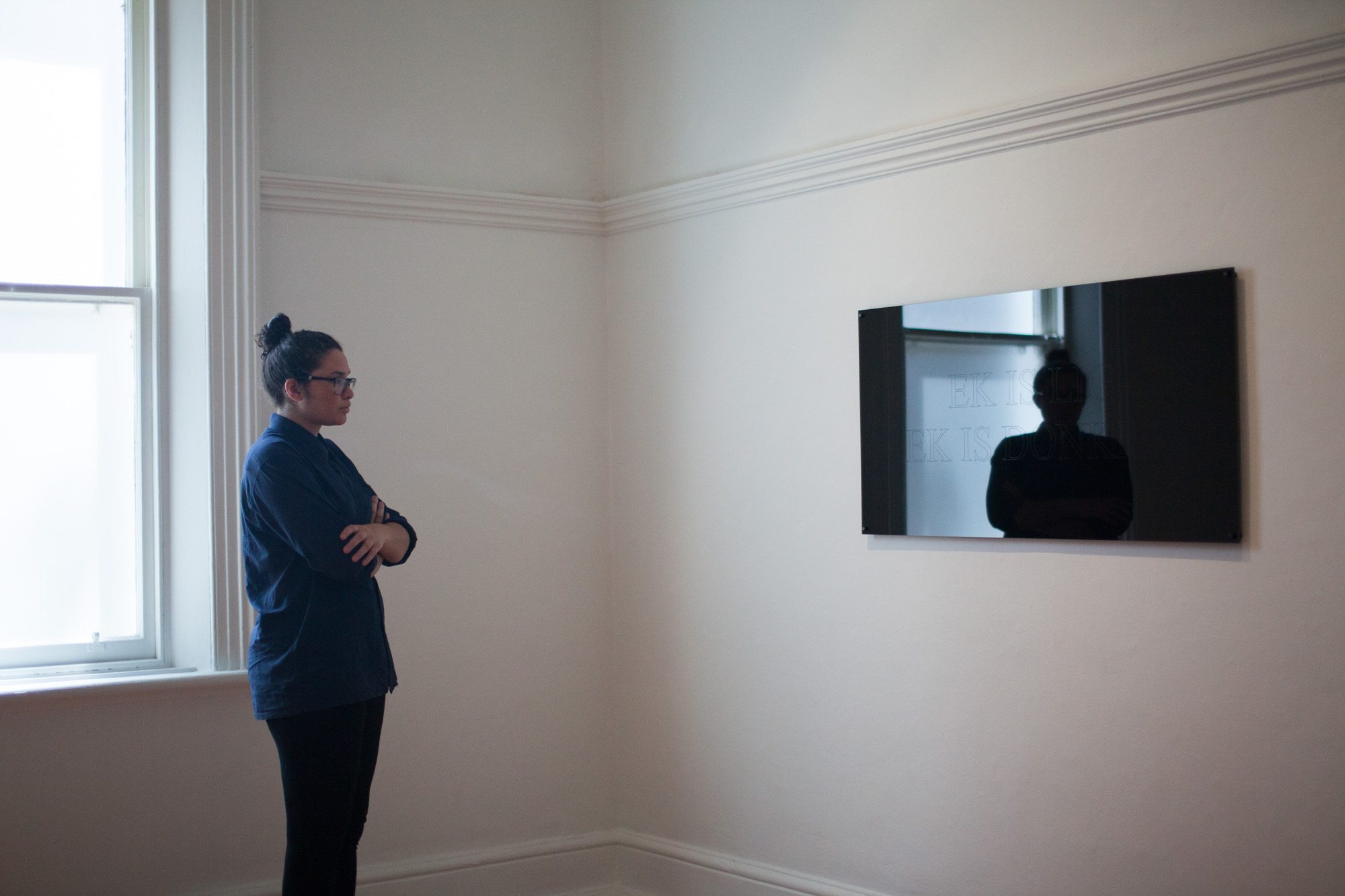 Installation view of a viewer with glasses and dark brown hair tied up, wearing a navy collar shirt and black pants, is standing before the mirrored work “EK IS LIG / EK IS DONKER,” their silhouette is reflected in its surface.