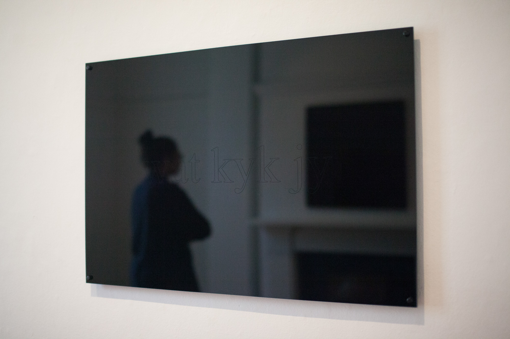 Angled view of the mirrored work “wat kyk jy,” reflecting a viewer standing with their arms folded, a fireplace, and another black perspex reflective artwork.