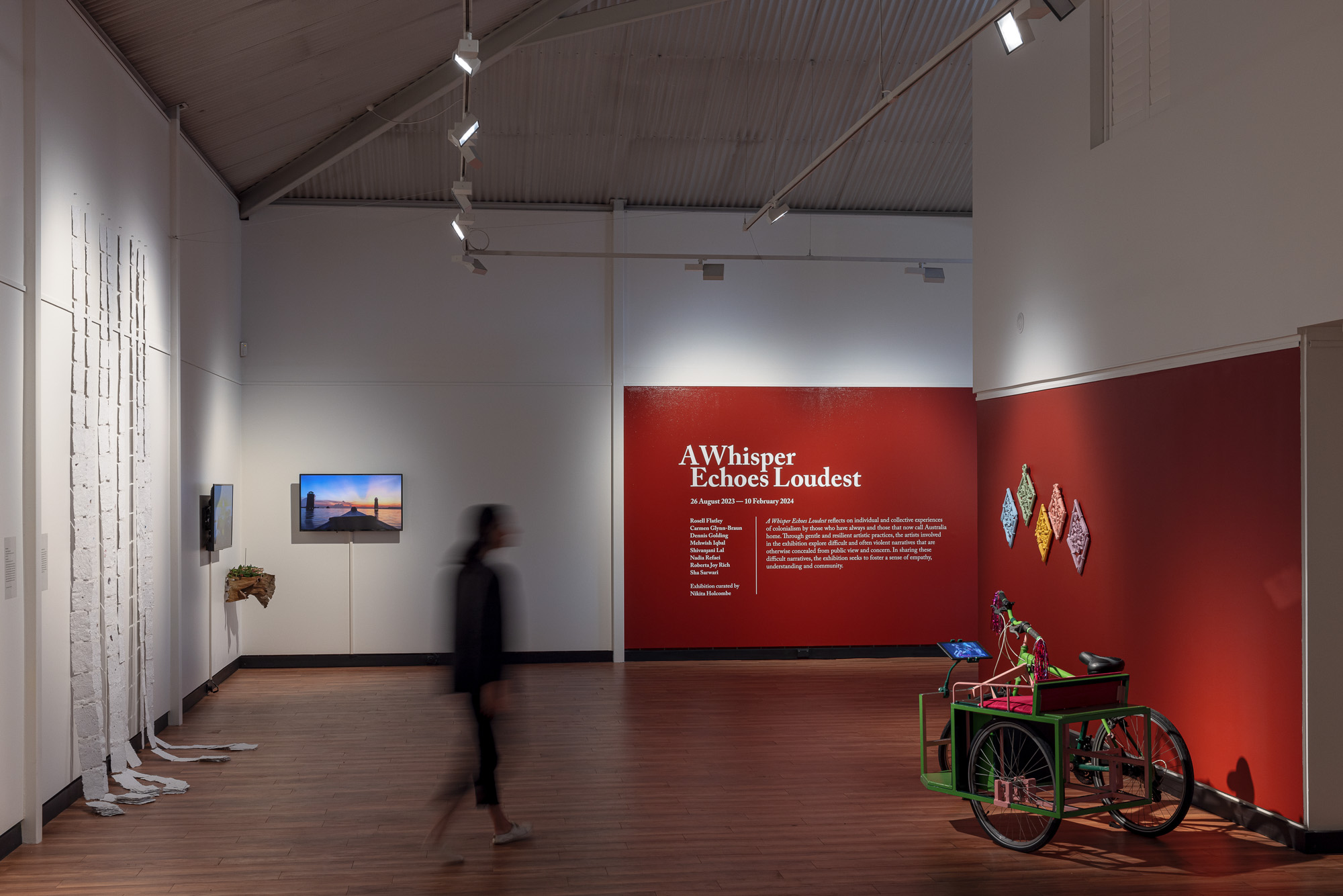 Installation view of 'A Whisper Echoes Loudest' in a high-ceilinged gallery. A blurred visitor crosses the wooden floor between a red exhibition wall, two video screens, a decorated tricycle, colourful relief sculptures, and a floor-to-ceiling white paper installation.