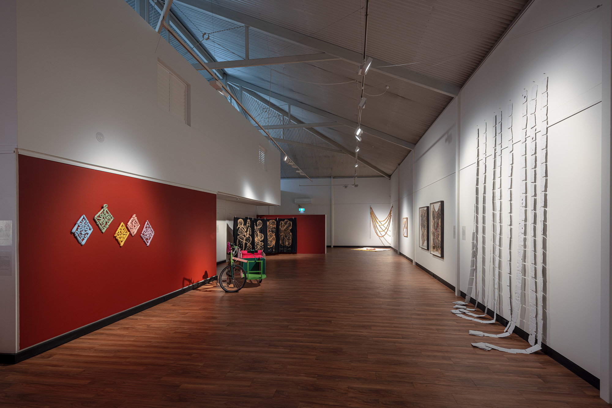 Wide view of 'A Whisper Echoes Loudest,' showing colourful relief sculptures and a decorated tricycle against a red wall, framed textiles and photography works throughout the gallery, and long white strips cascading down the right wall.