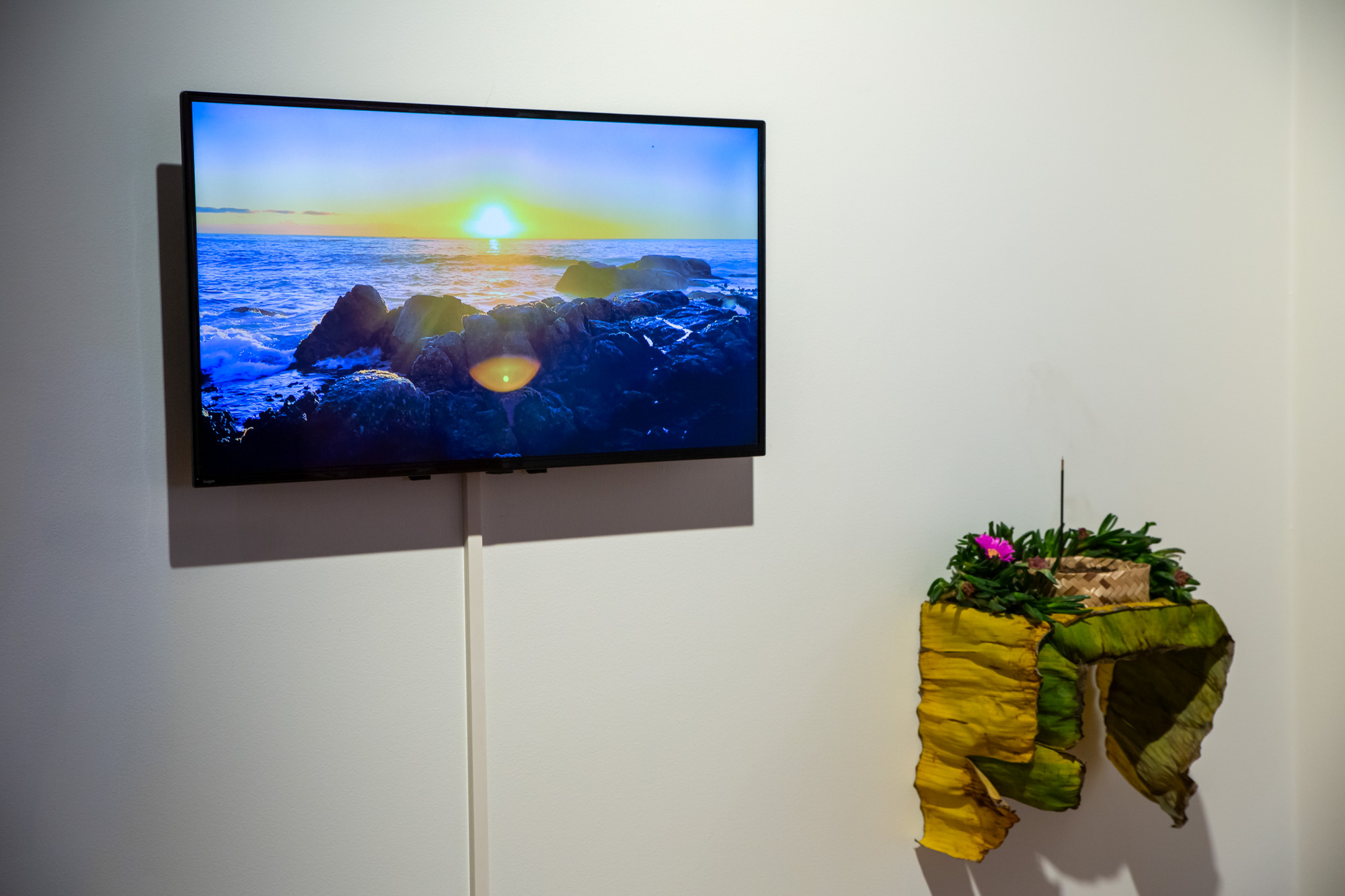 Installation detail showing a video of the sun setting over a rocky ocean shoreline on the Atlantic seaboard in Cape Town, beside a wall-mounted installation made from organic material including yellow and green banana leaves, pink flowers, soil, a woven bamboo basket, and incense.