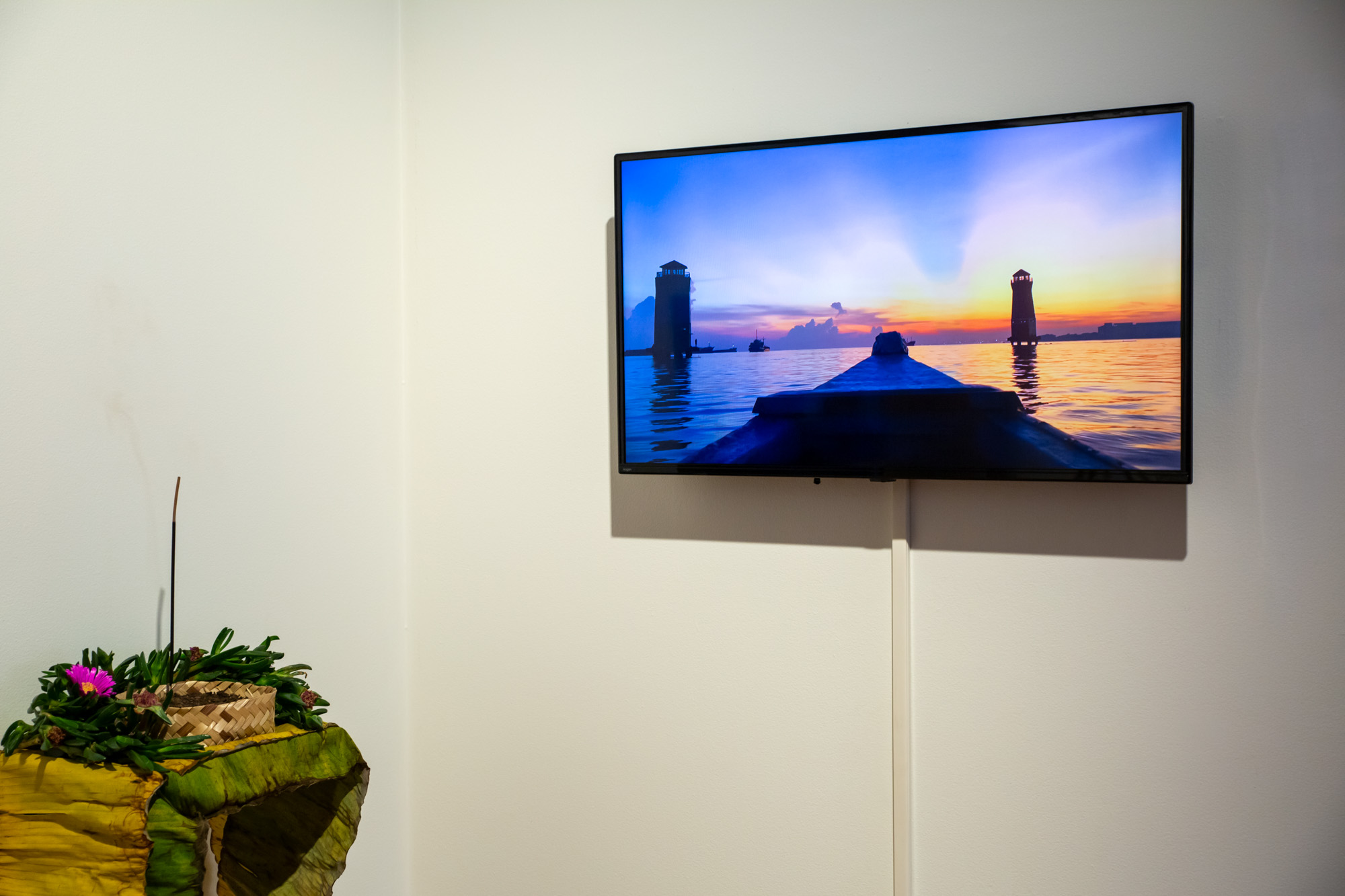 Installation detail showing a video filmed from the bow of a wooden boat floating at Sunda Kelapa port approaching two tower-like structures at sunrise, displayed beside a wall-mounted installation made from organic material including yellow and green banana leaves, pink flowers, soil, a woven bamboo basket, and incense.