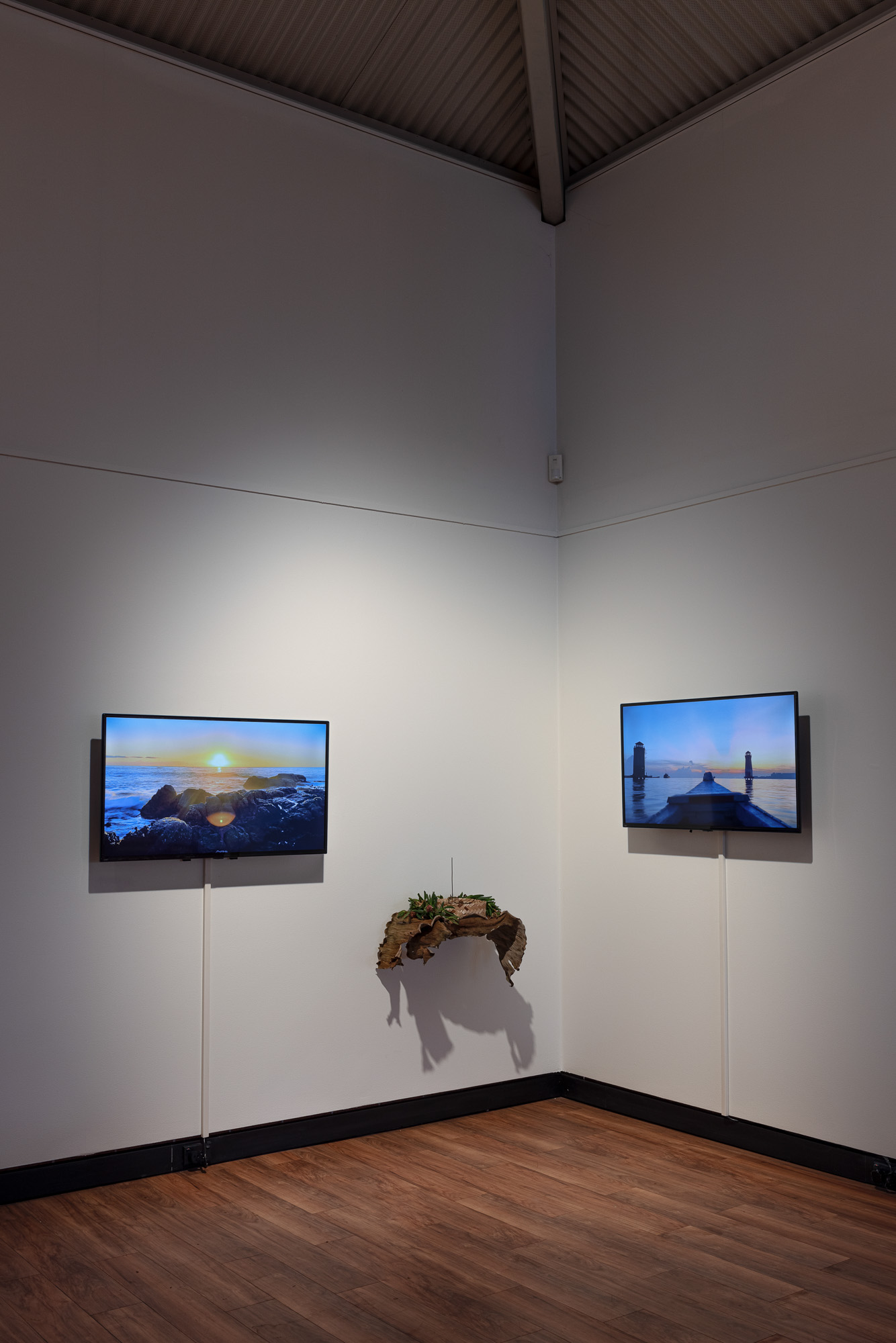 Corner installation view of two video screens on adjoining walls: one shows a sunset over a rocky shoreline, while the other shows a boat floating at sunrise approaching two towers. An organic sculpture of dried leaves, plants, soil, and woven material hangs between them.