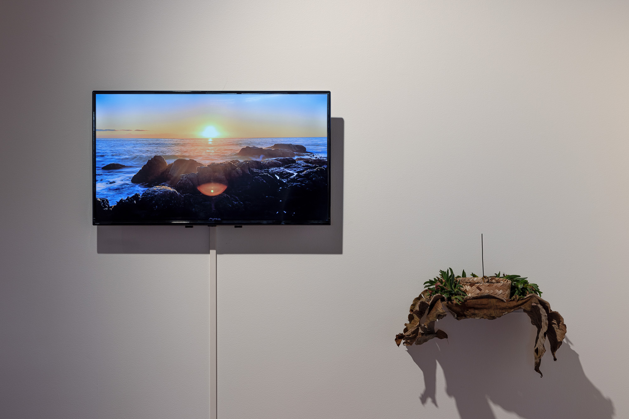 Installation detail of a video showing the sun setting low over a rocky ocean shoreline, displayed beside an organic wall sculpture made from dried leaves, green foliage, soil, woven bamboo basket holding an upright incense stick.