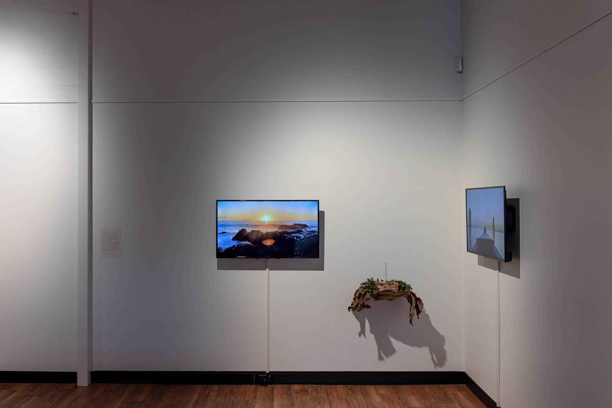 Wide installation view of two video screens on adjacent gallery walls, one showing a rocky shoreline at sunset and the other a boat floating at sunrise moving toward two port towers. Between them hangs an organic sculpture casting an elongated shadow.