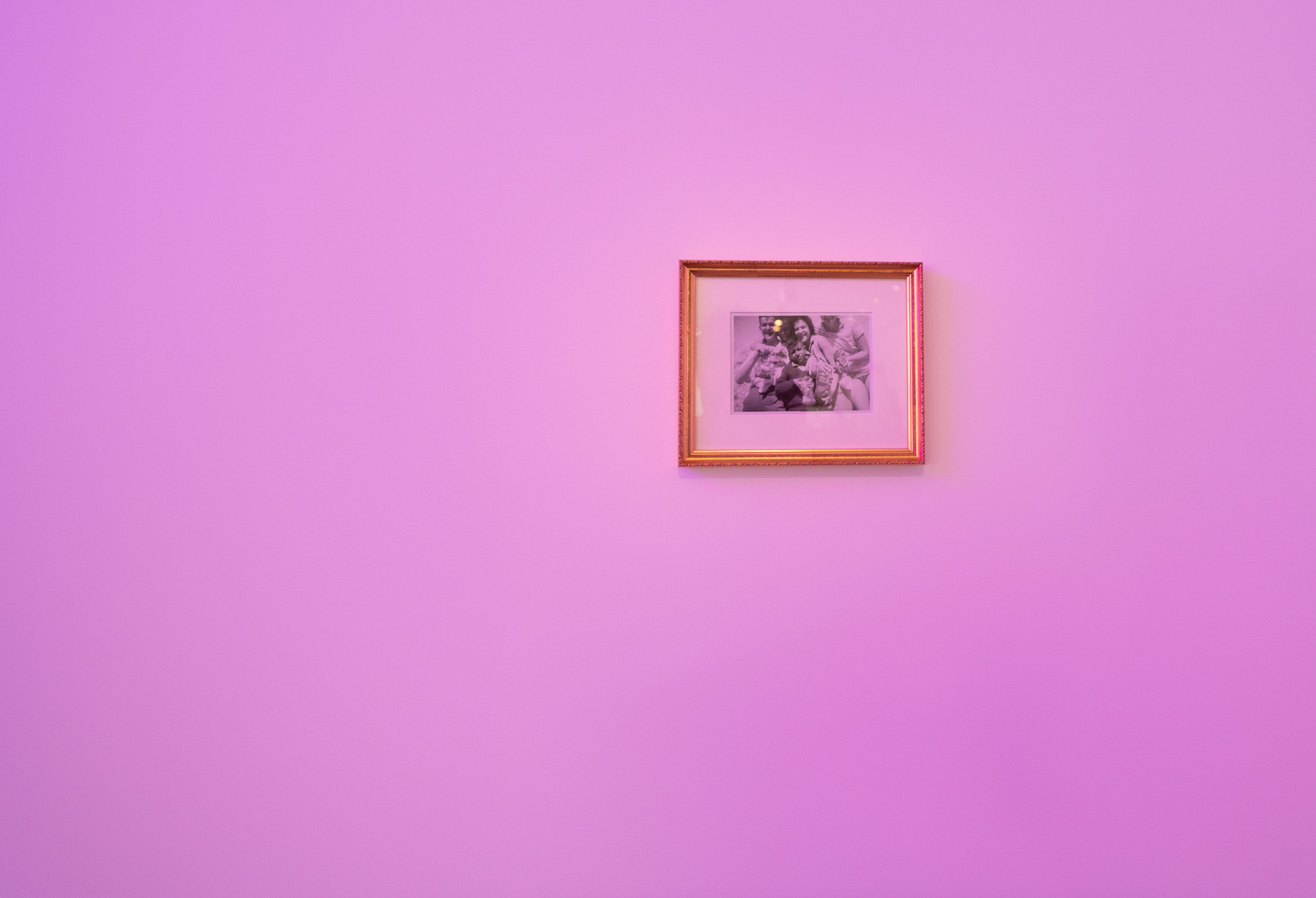 A small black-and-white family photograph in an ornate gold frame, showing a group of adults and children gathered closely together, displayed on a wall washed in pink light.