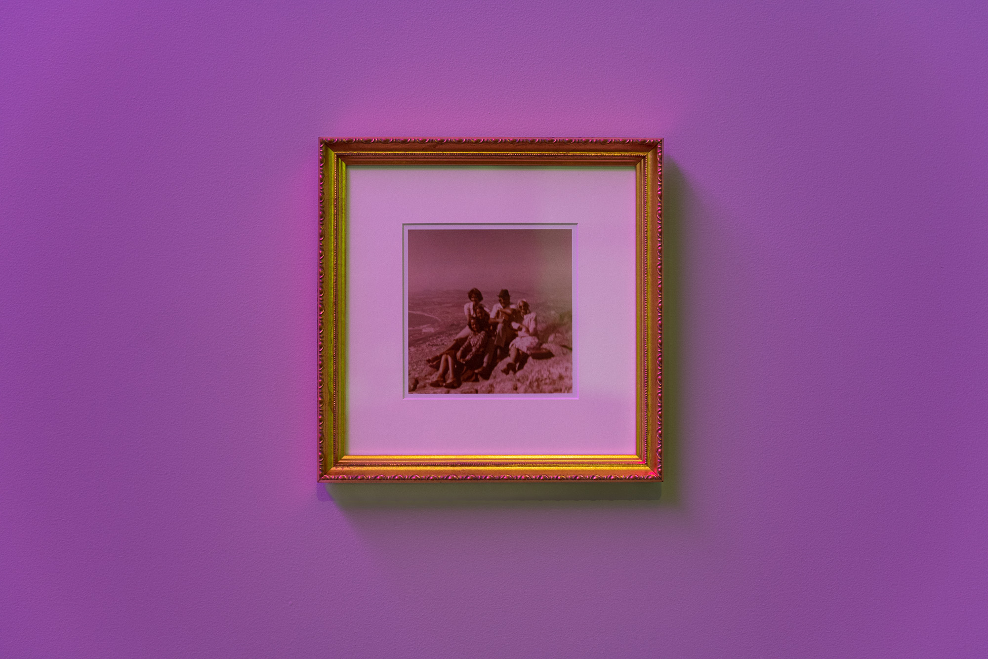 Close view of an ornate gold frame containing a archival family photograph of several African people seated together on a hillside (Signal Hill, Cape Town), surrounded by a wide white mount and purple gallery lighting.
