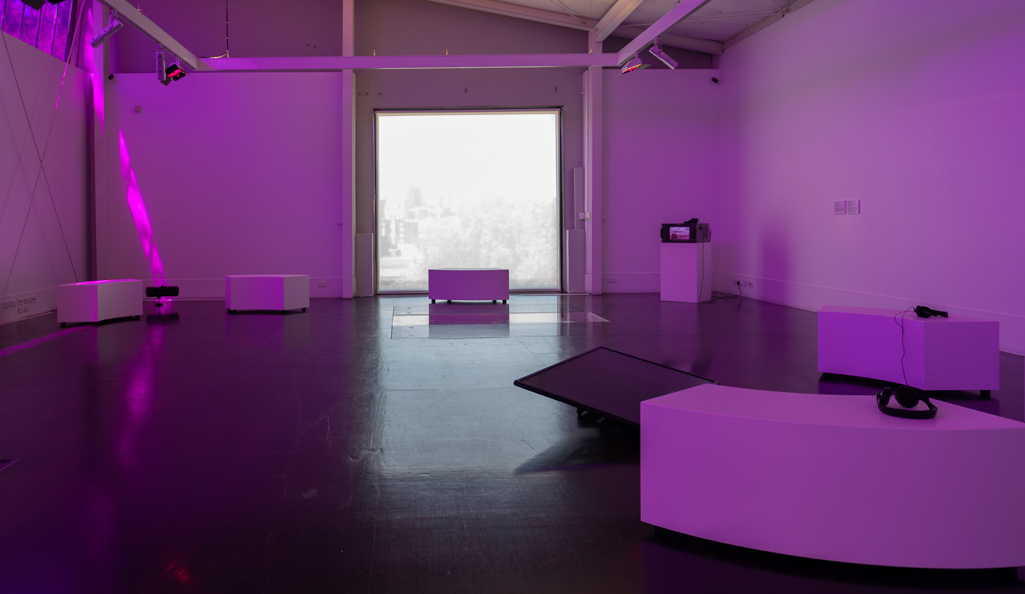 Wide installation view of a darkened gallery bathed in purple light, with curved white seating, headphones, video equipment, and a glass-covered opening in the floor arranged around a large luminous window.