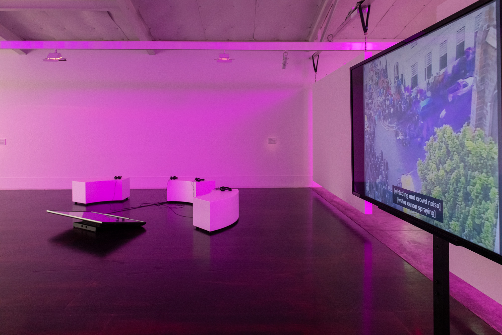 Multimedia installation view with a large flatscreen monitor on a tall stand showing aerial footage of a crowded street and subtitles reading “[whistling and crowd noise] [water canon spraying].” Curved seats with headphones and a low screen occupy the purple-lit gallery.