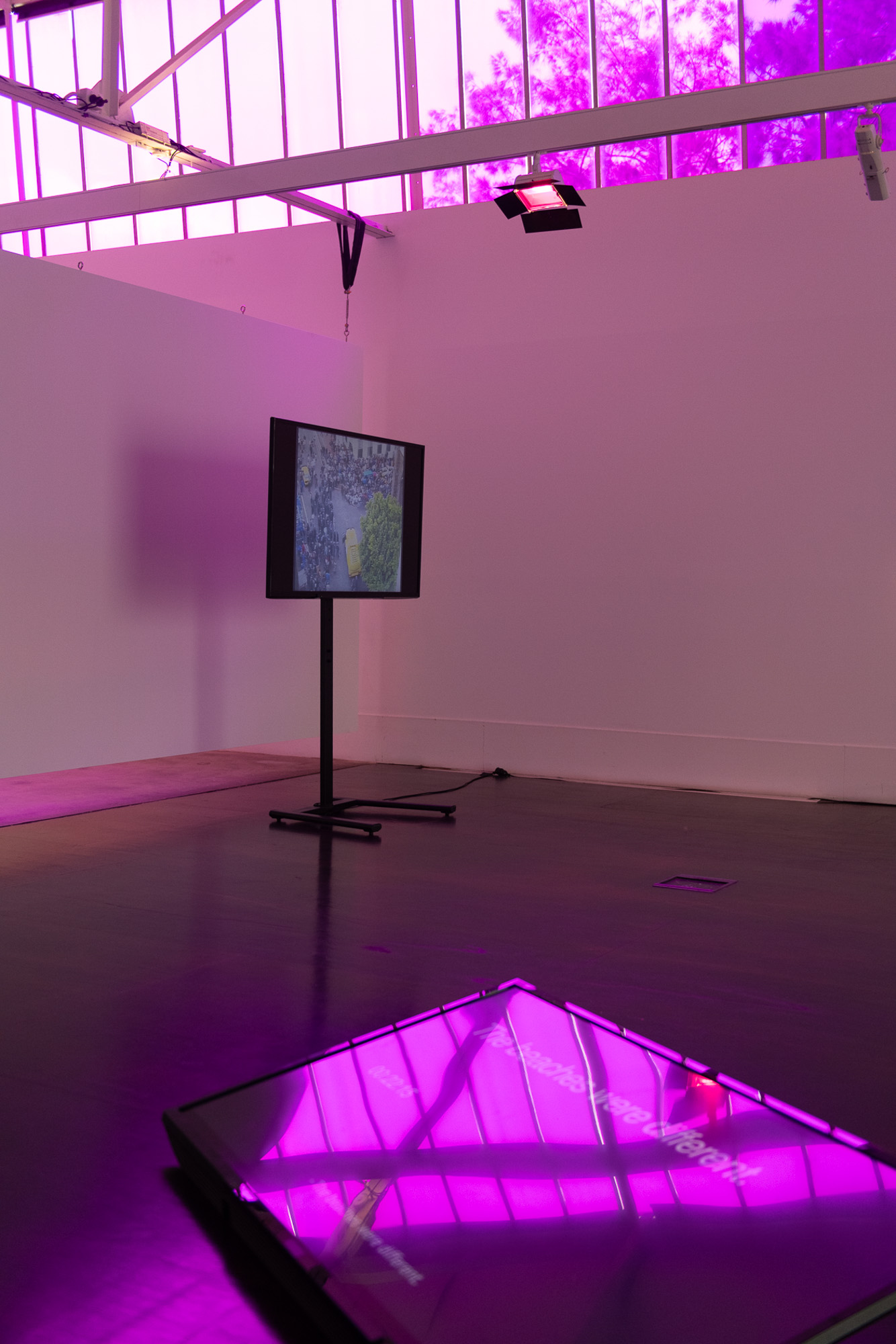 Installation detail showing a freestanding monitor playing aerial footage of a crowd, with a large horizontal screen lying near the floor beneath purple-filtered windows and theatrical lighting.